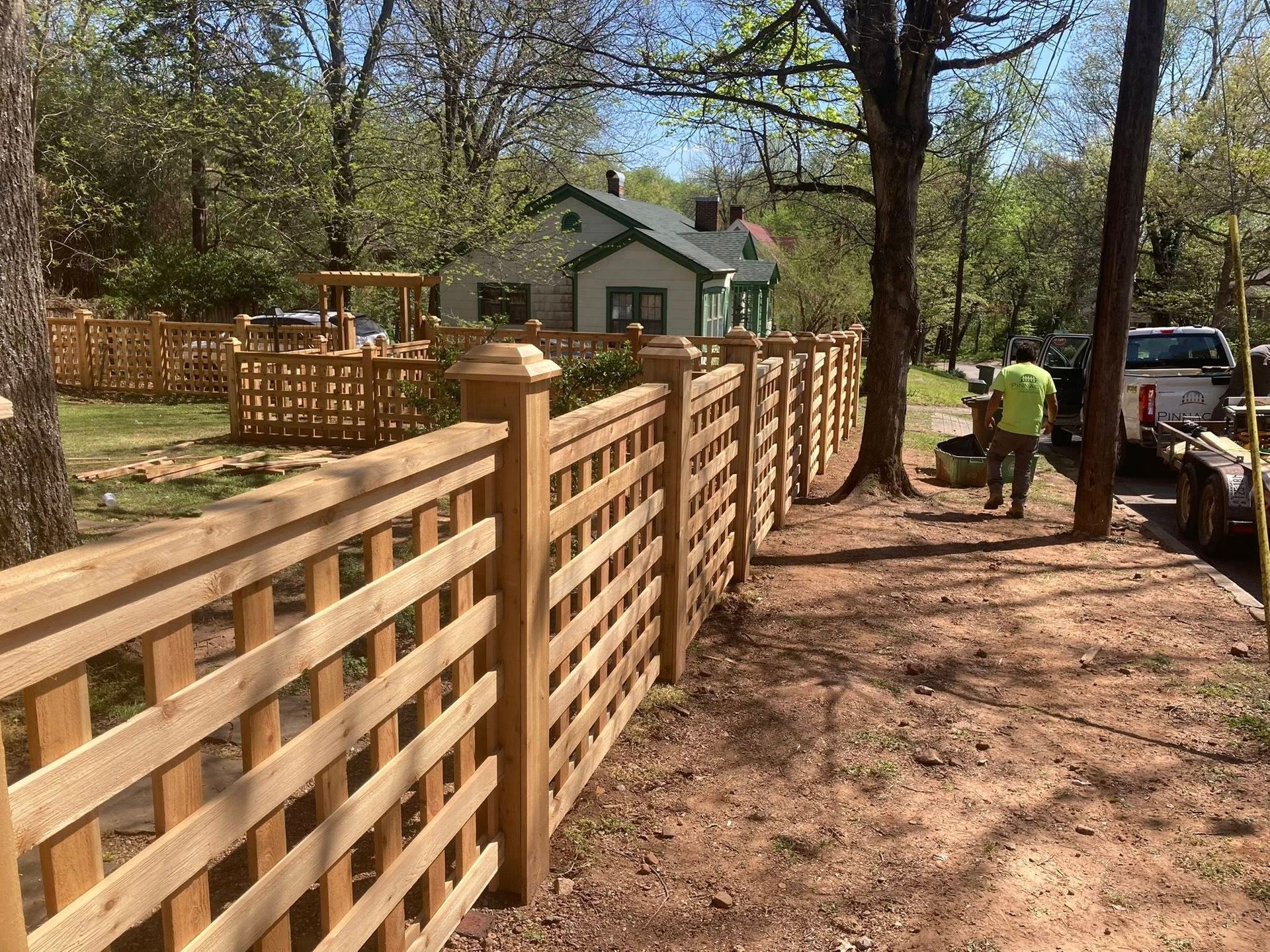 Wooden fence along a dirt path with workers and a white truck in a wooded yard