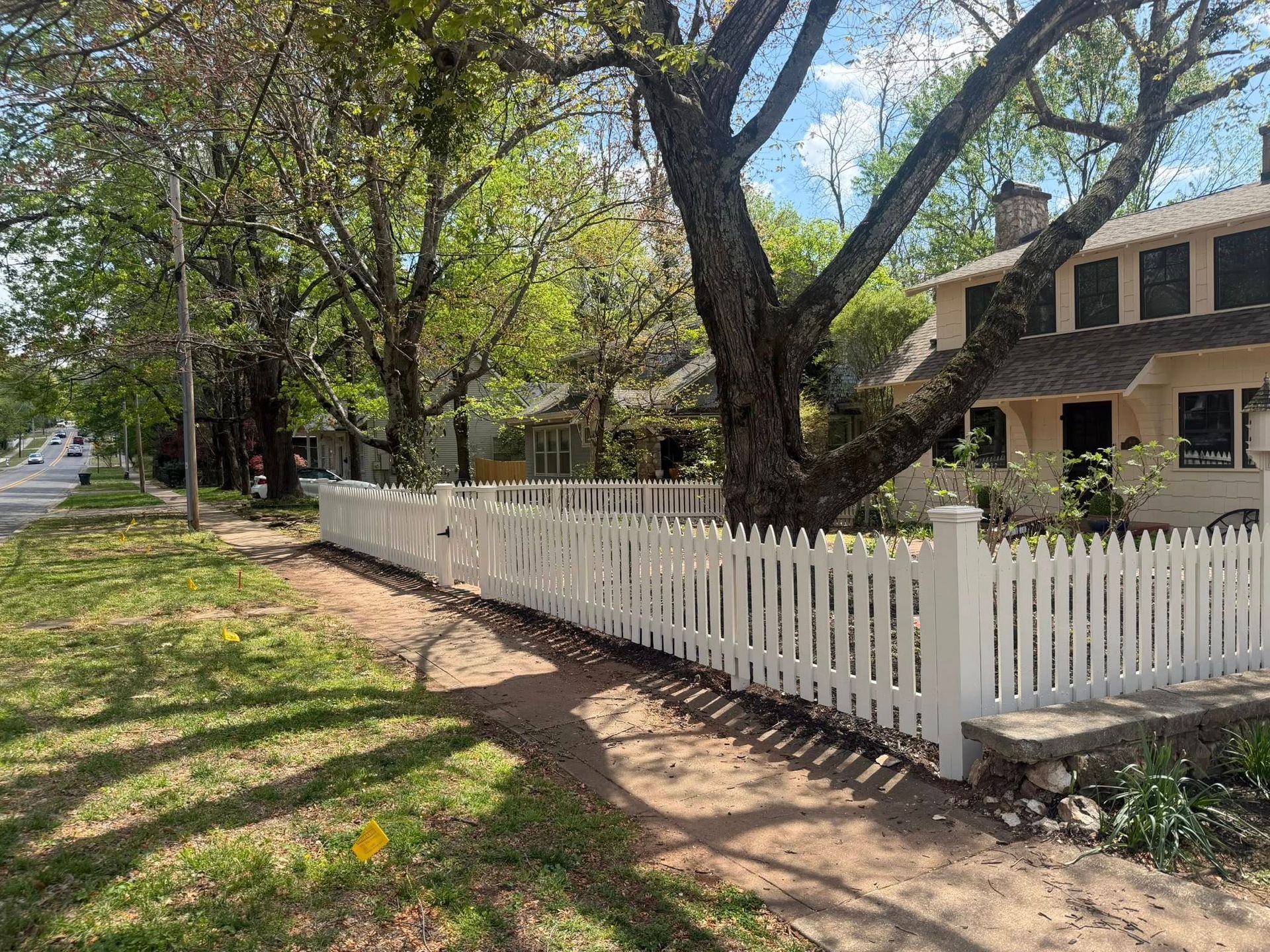 White picket fence along a tree-lined suburban sidewalk beside houses on a sunny day