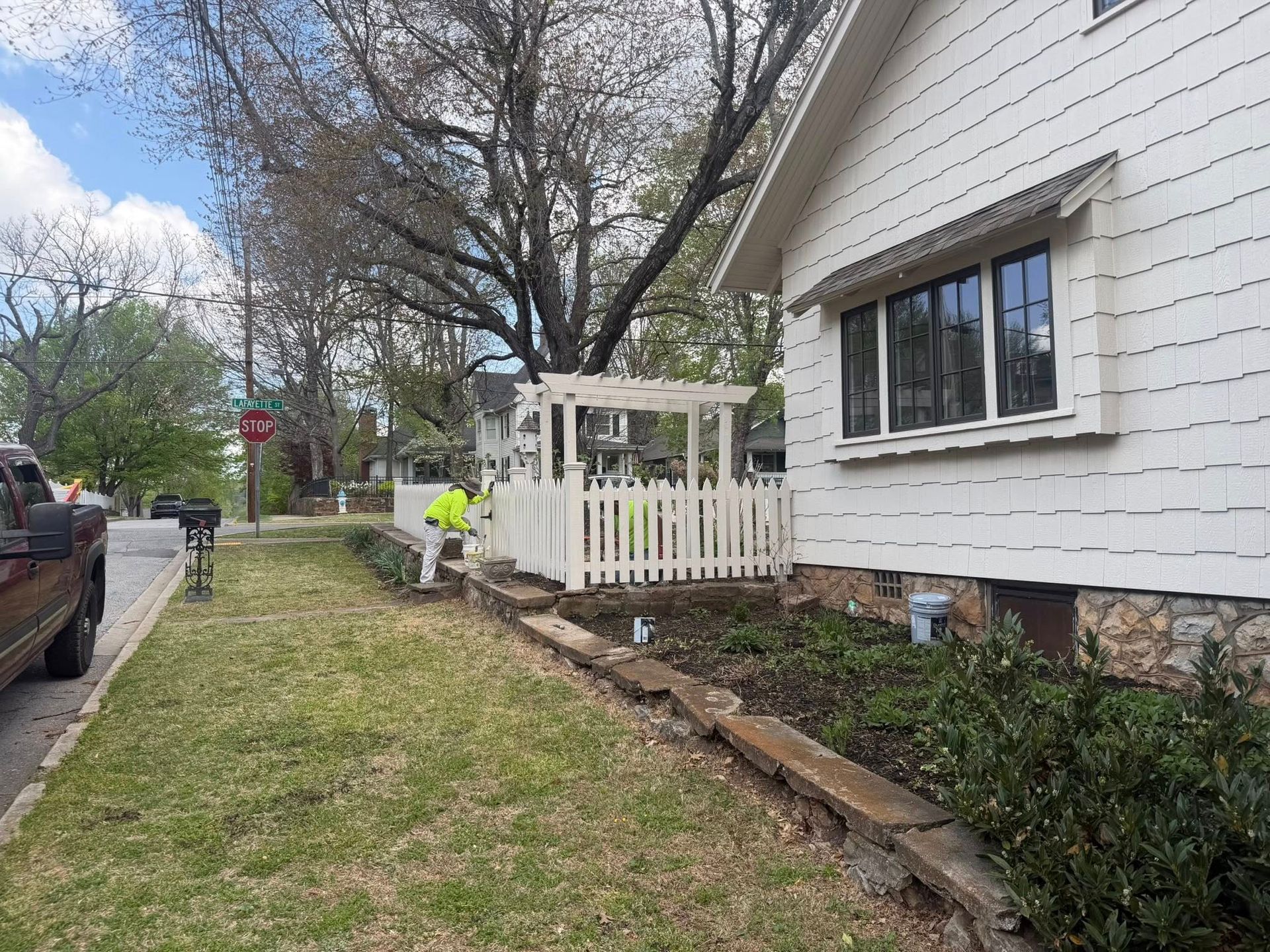 Worker in neon vest clearing debris along a white house’s side yard beside a sidewalk and parked car