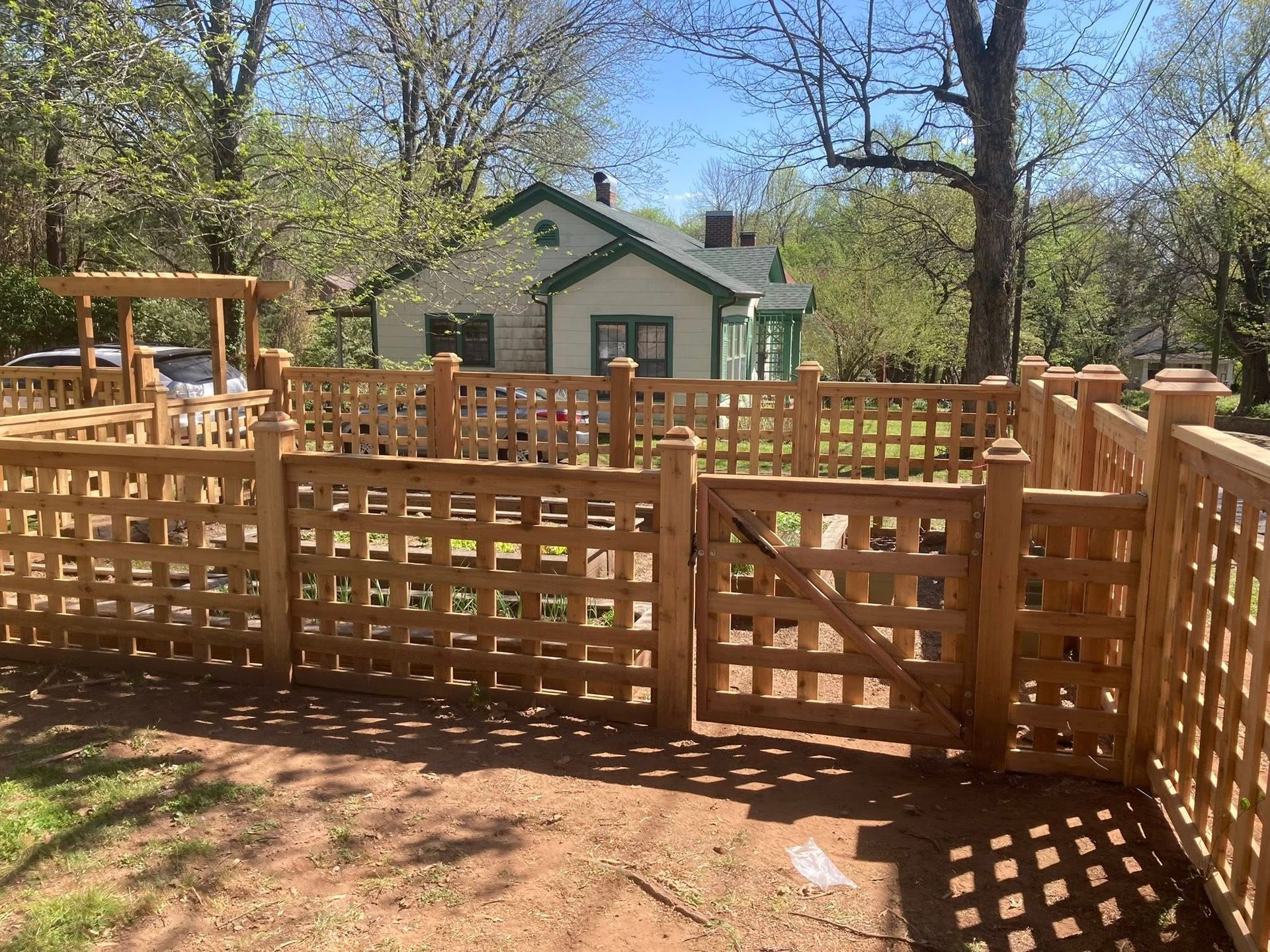 Wooden fenced deck with gate in a wooded yard, with a small house in the background.