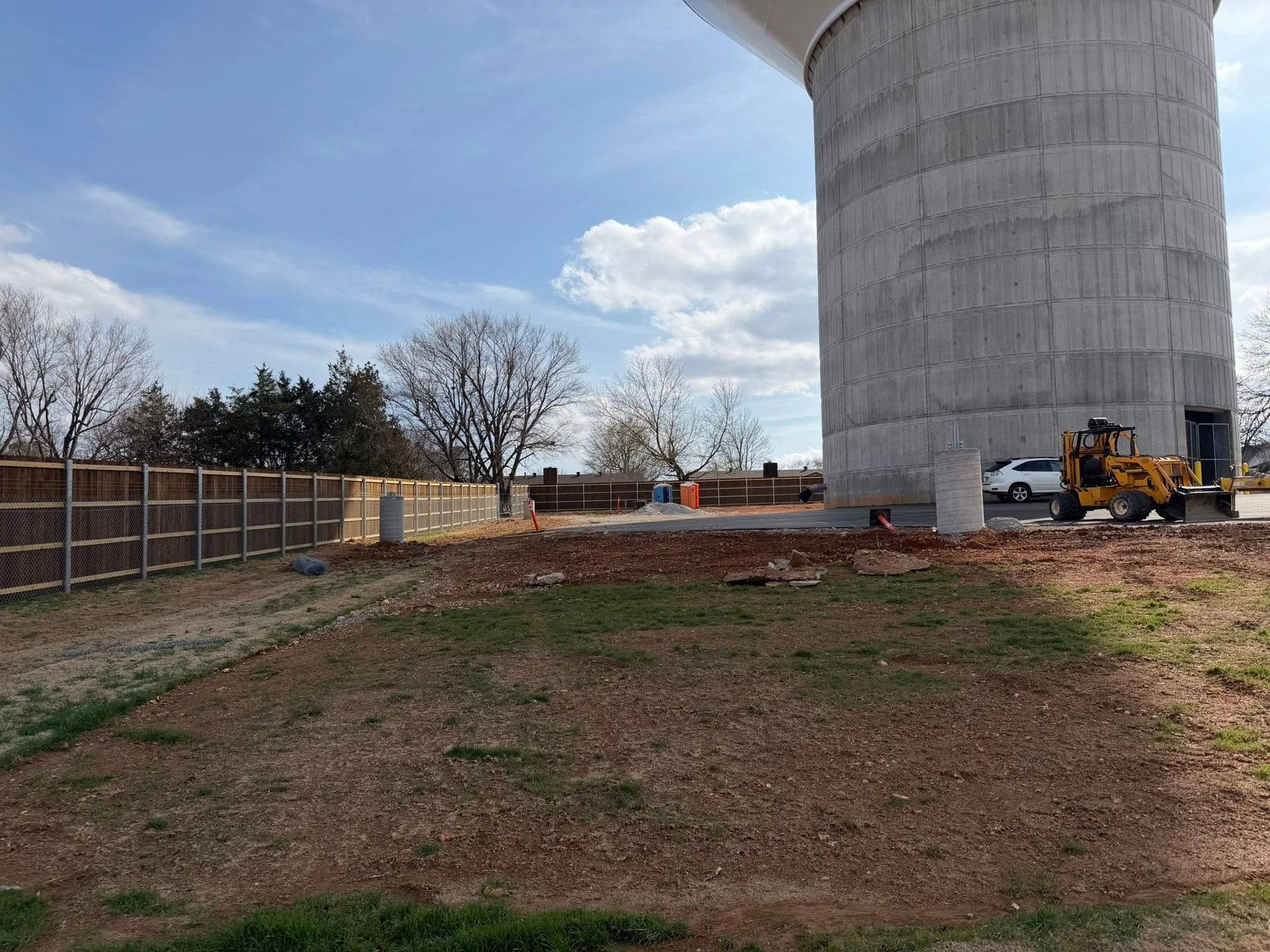 Construction site beside a large concrete tower, with dirt foreground, fence, and a yellow loader.