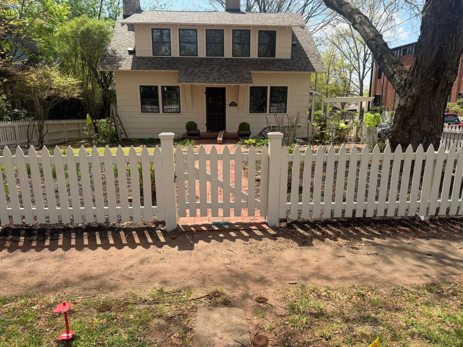 Two-story beige house behind a white picket fence, with a large tree in the front yard.