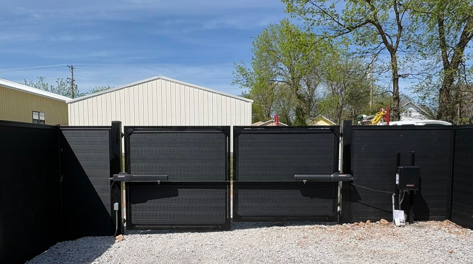 Black metal gate panels beside a gravel yard, with a white building and trees under a blue sky.