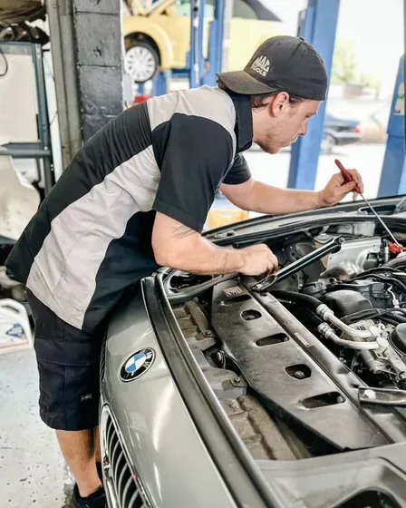 Mechanic working on a car engine in a garage; wearing a cap, shirt, and shorts.