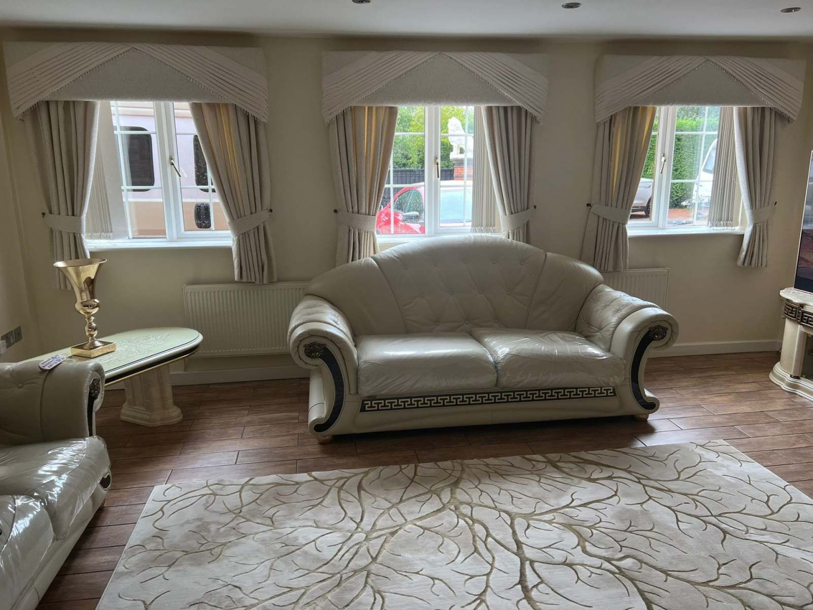 Elegant living room with cream sofas, patterned rug, and three curtained windows overlooking a street.