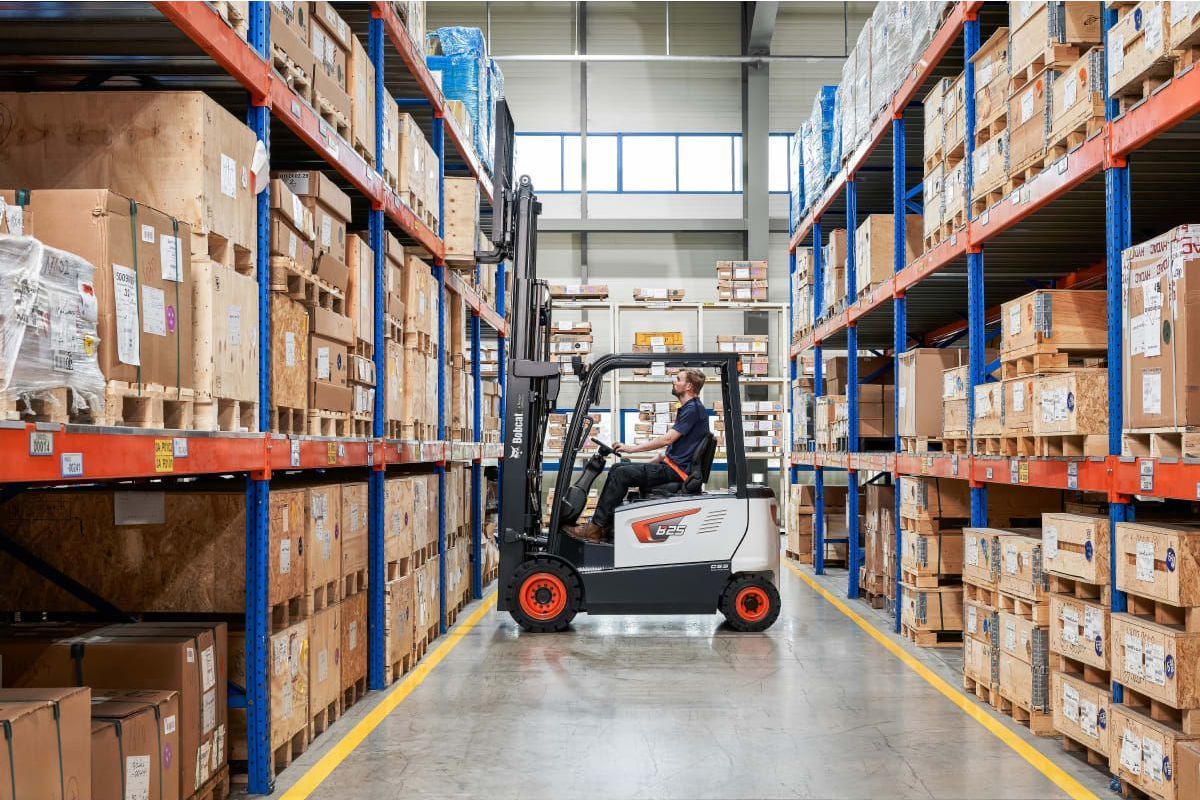 A man is driving a forklift in a warehouse filled with lots of boxes.
