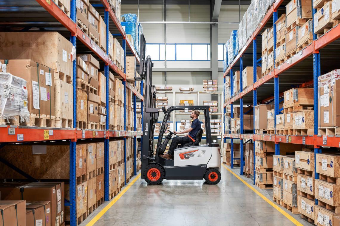 A man is driving a forklift in a warehouse filled with lots of boxes.