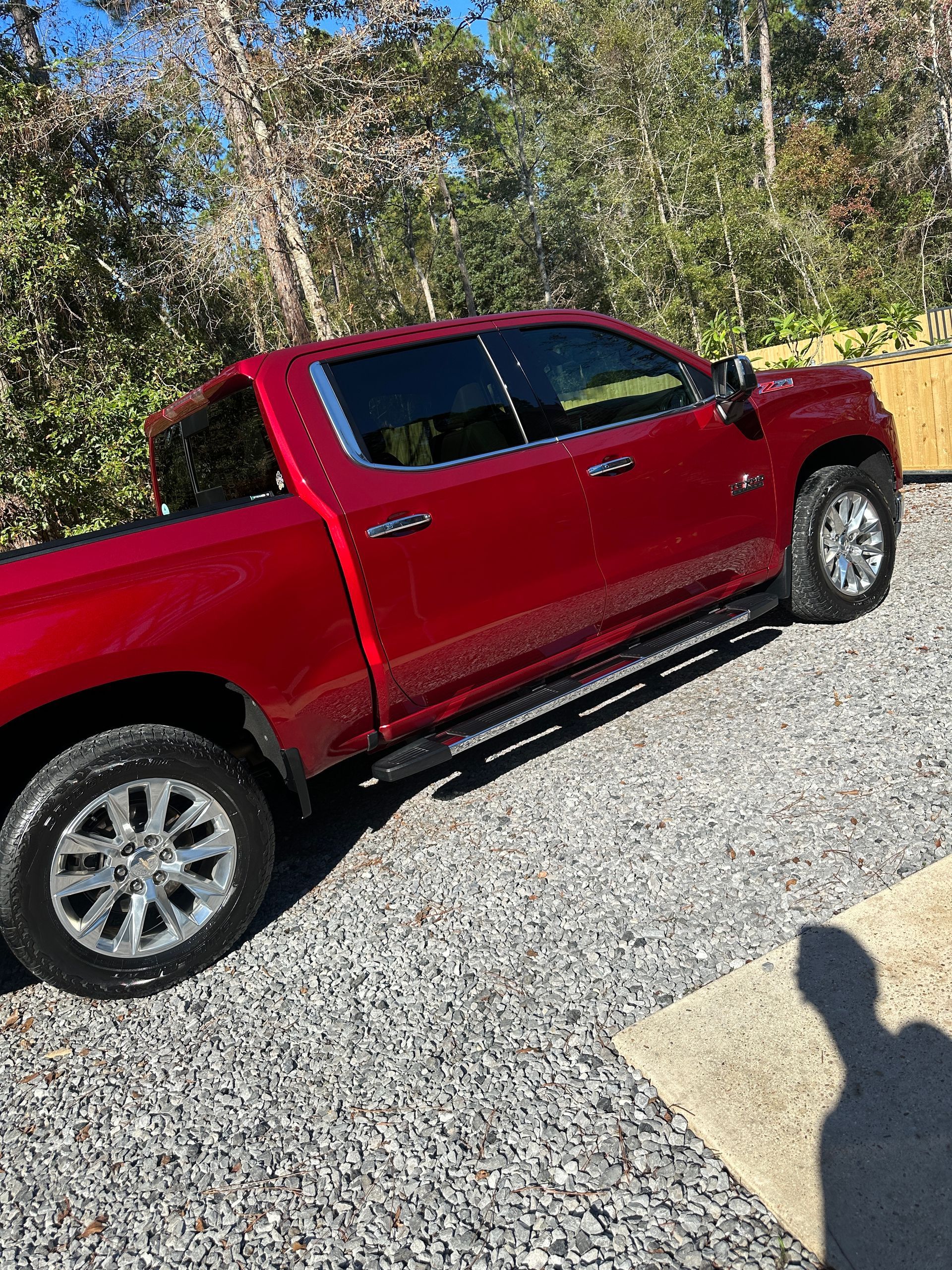 Red pickup truck parked on a gravel driveway beside trees in bright sunlight.