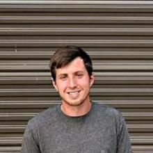 Smiling man in a gray shirt standing in front of a brown metal shutter