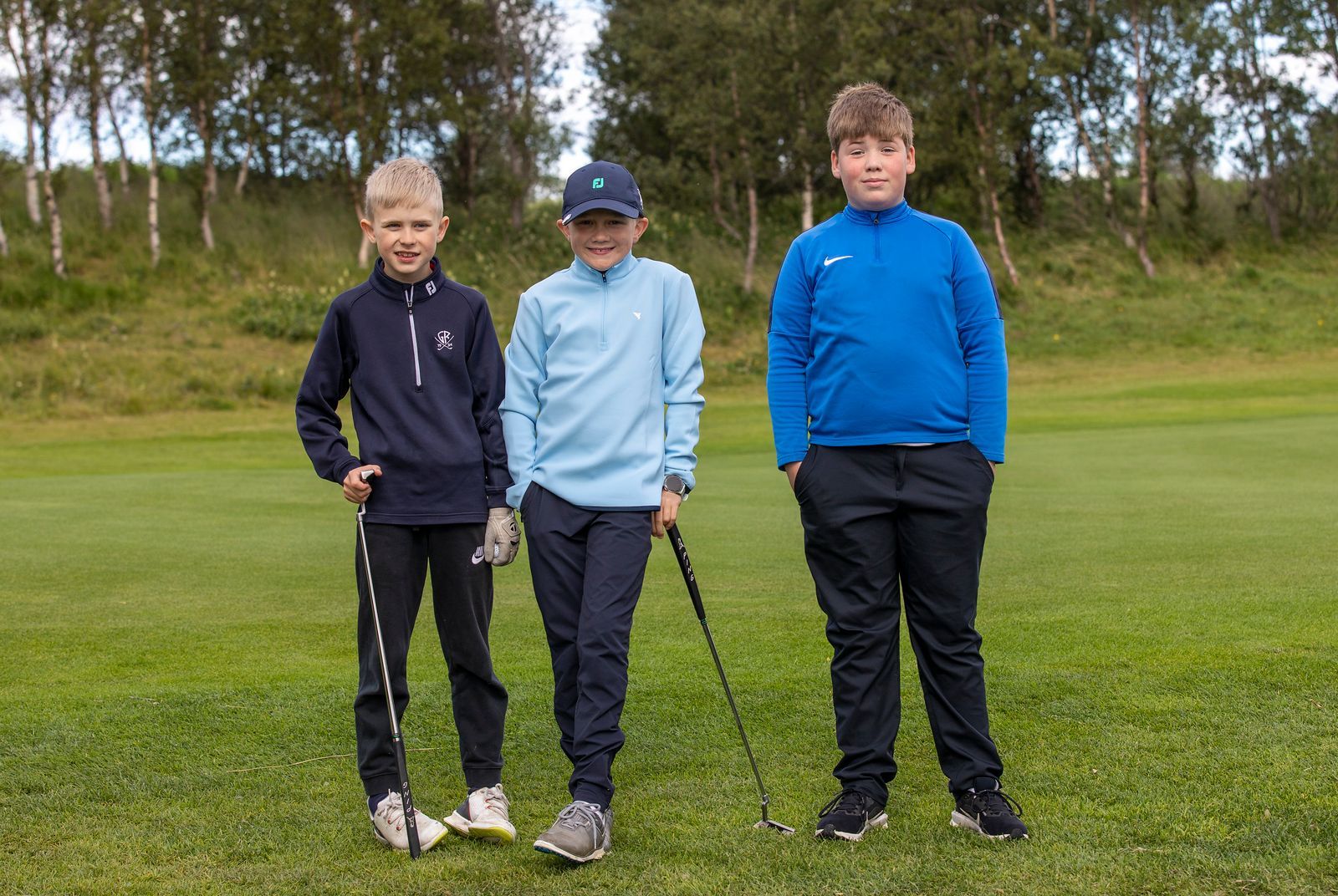 Þrjú börn standa á grasvelli með golfkylfur í höndunum, með tré í bakgrunni.