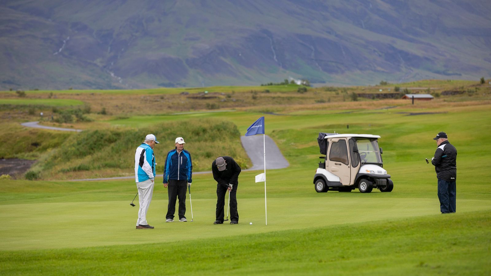 Golfarar við púttvöll nálægt fána og golfbíl, með fjöllum í bakgrunni.