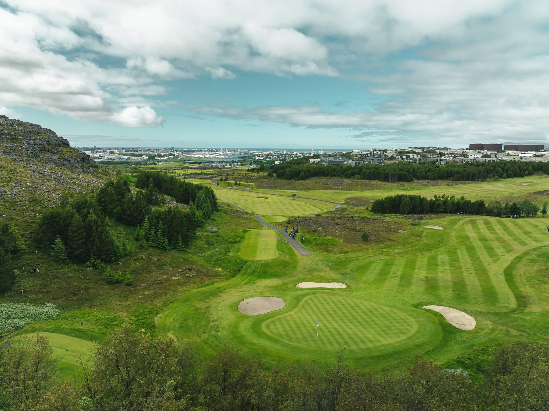 Grænn golfvöllur með sandbyrgjum, öldóttum brautum og fjarlægum hæðum undir skýjuðum himni