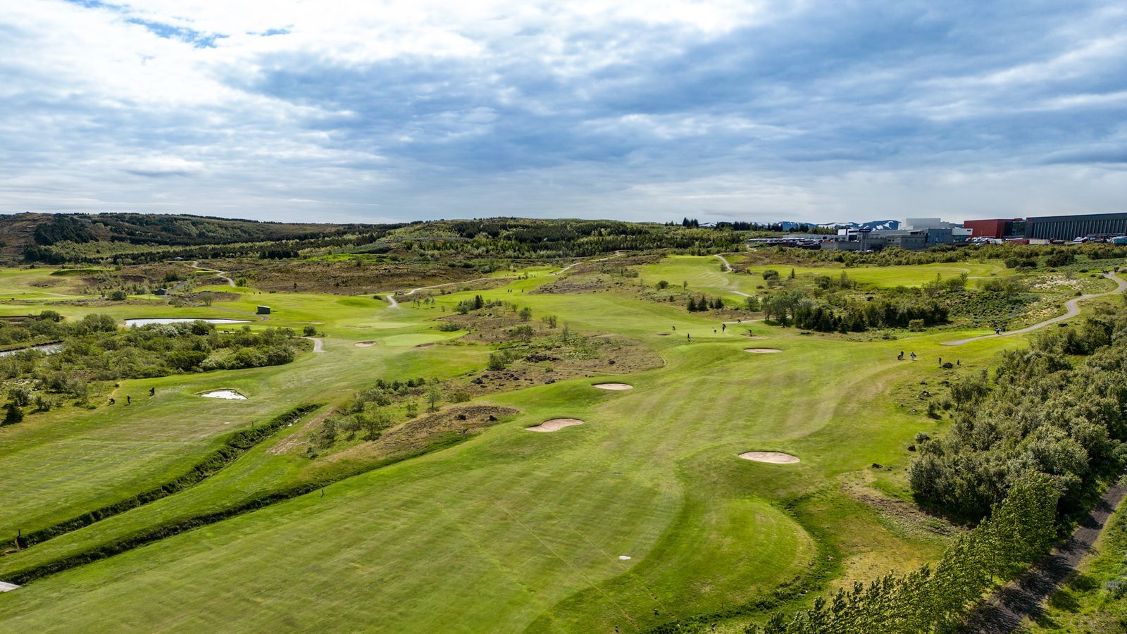 Grænn golfvöllur með öldóttum brautum, sandbyrgjum og skýjuðum himni í bakgrunni