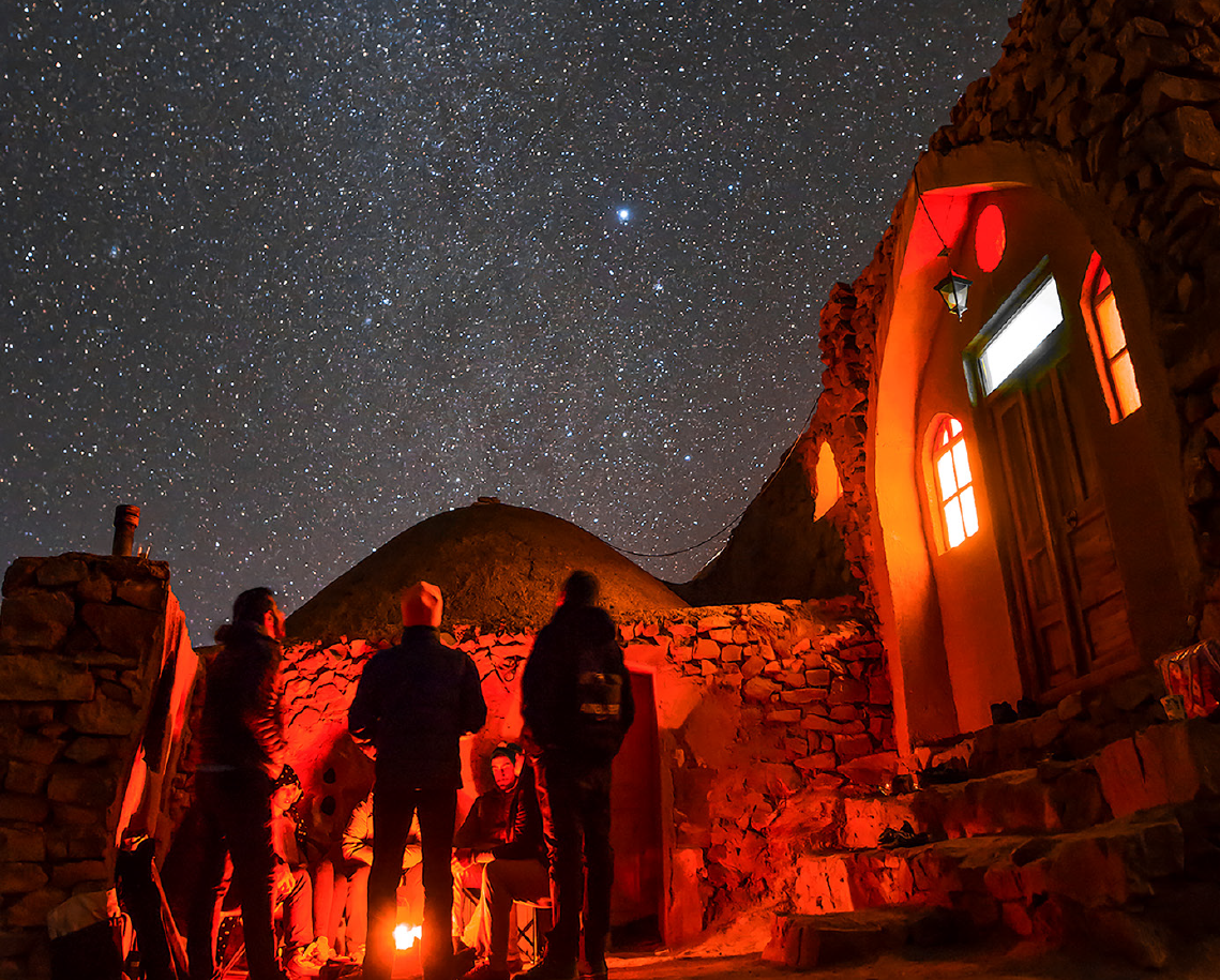 Three people gazing at a starry night sky near a stone building with warm interior lighting.