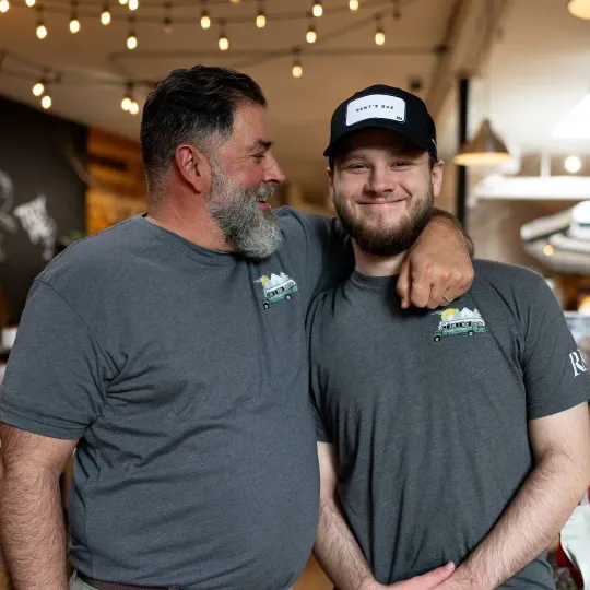 Two men in matching gray shirts smiling with arms around each other in a warmly lit indoor setting