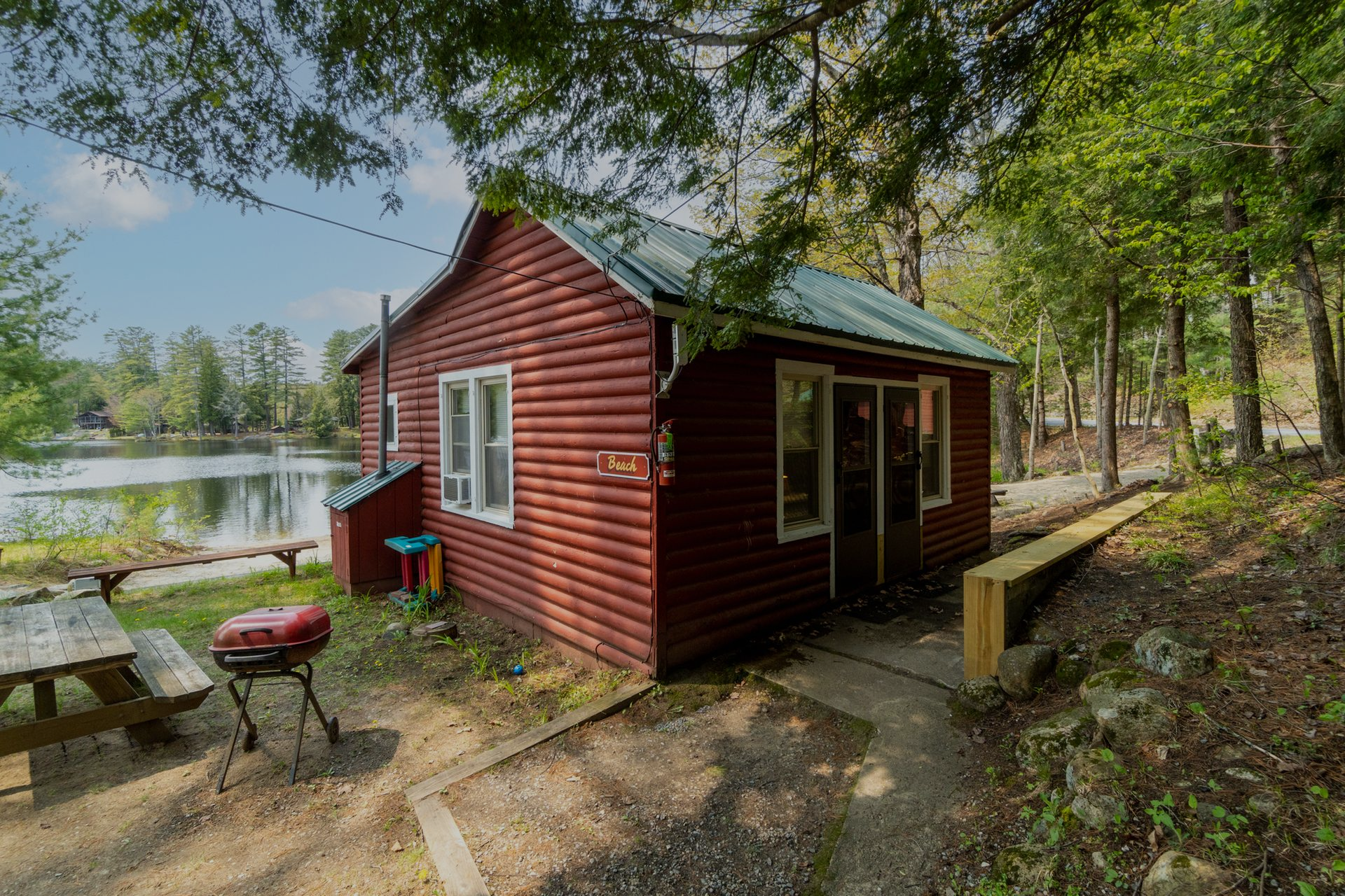 Red cabin by a lake with a picnic table and grill. Green trees surround the cabin.