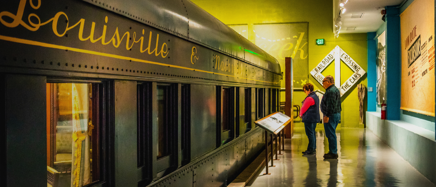 Visitors looking at a train car exhibit in a museum. The train car is labeled