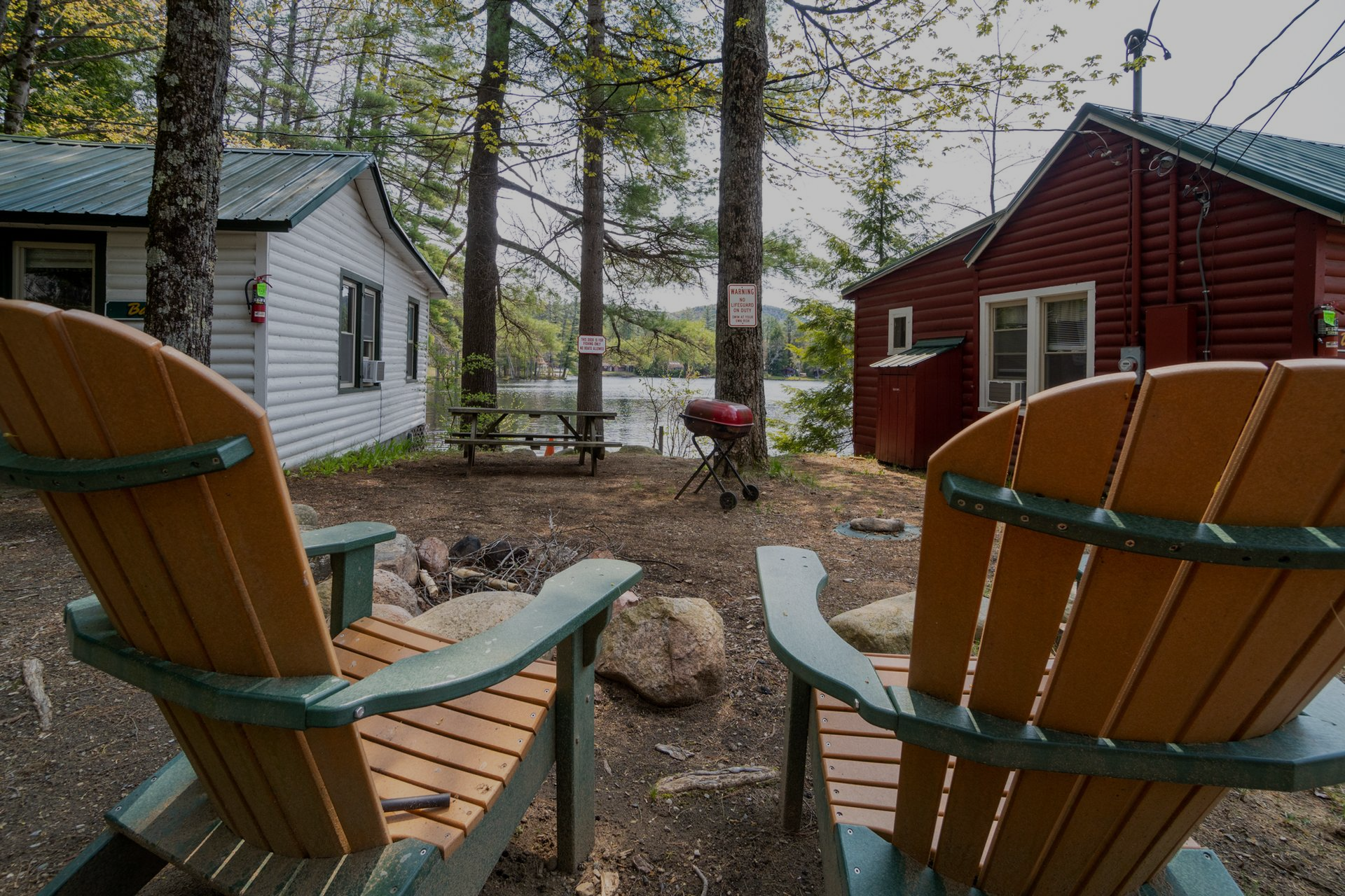 Two wooden Adirondack chairs face a lake, with cabins and a grill in the background.
