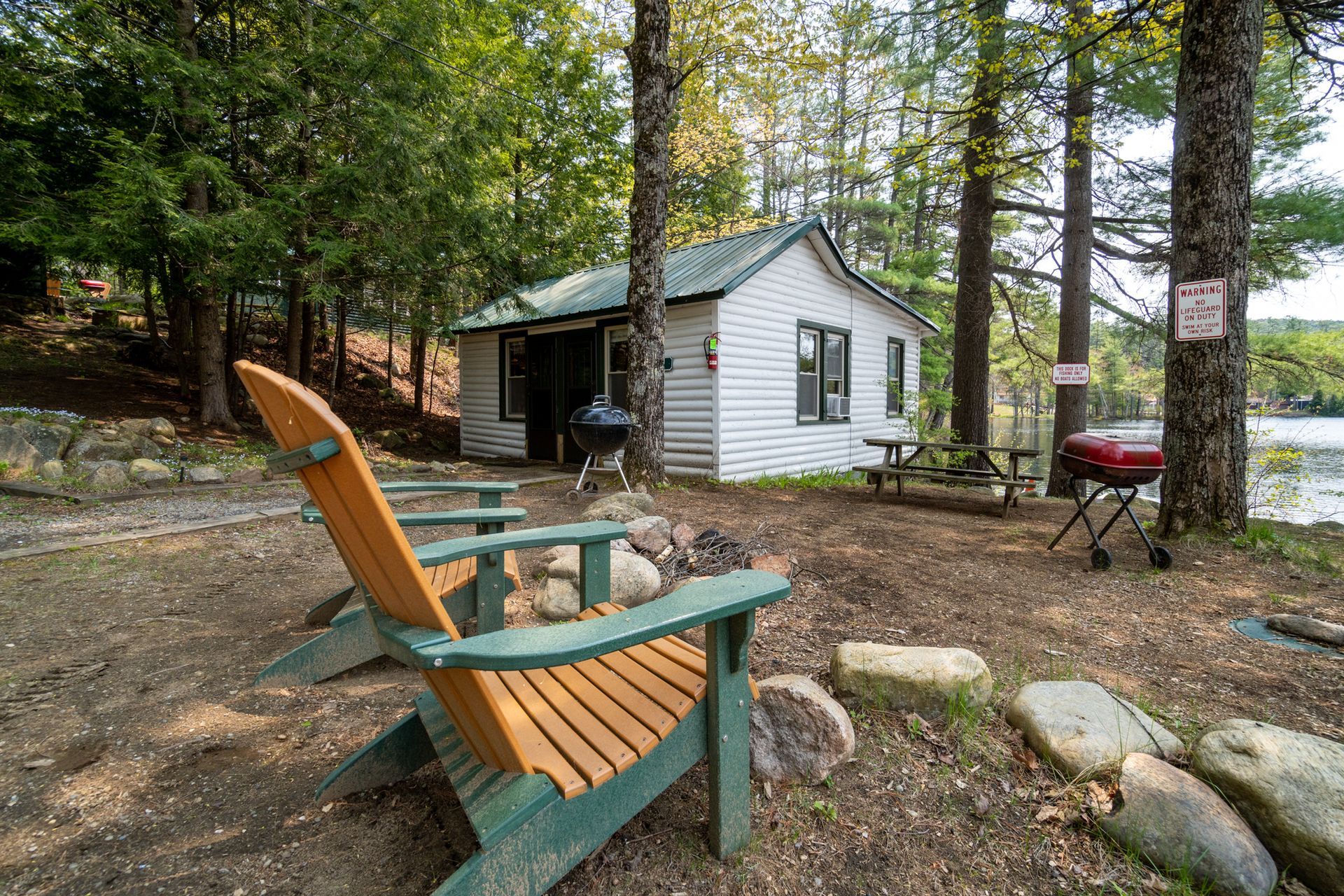 Two Adirondack chairs face a small lakeside cabin nestled among trees.