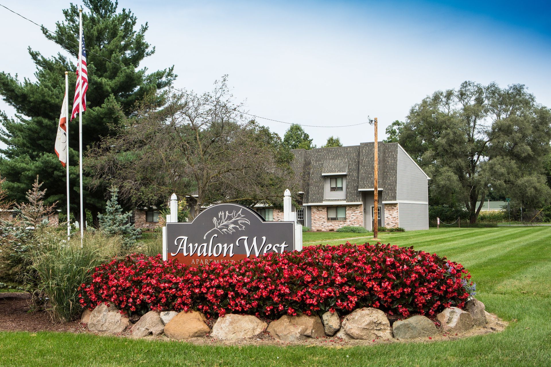 Avalon West sign in front of red flowers, lawn, and a building with an American flag.