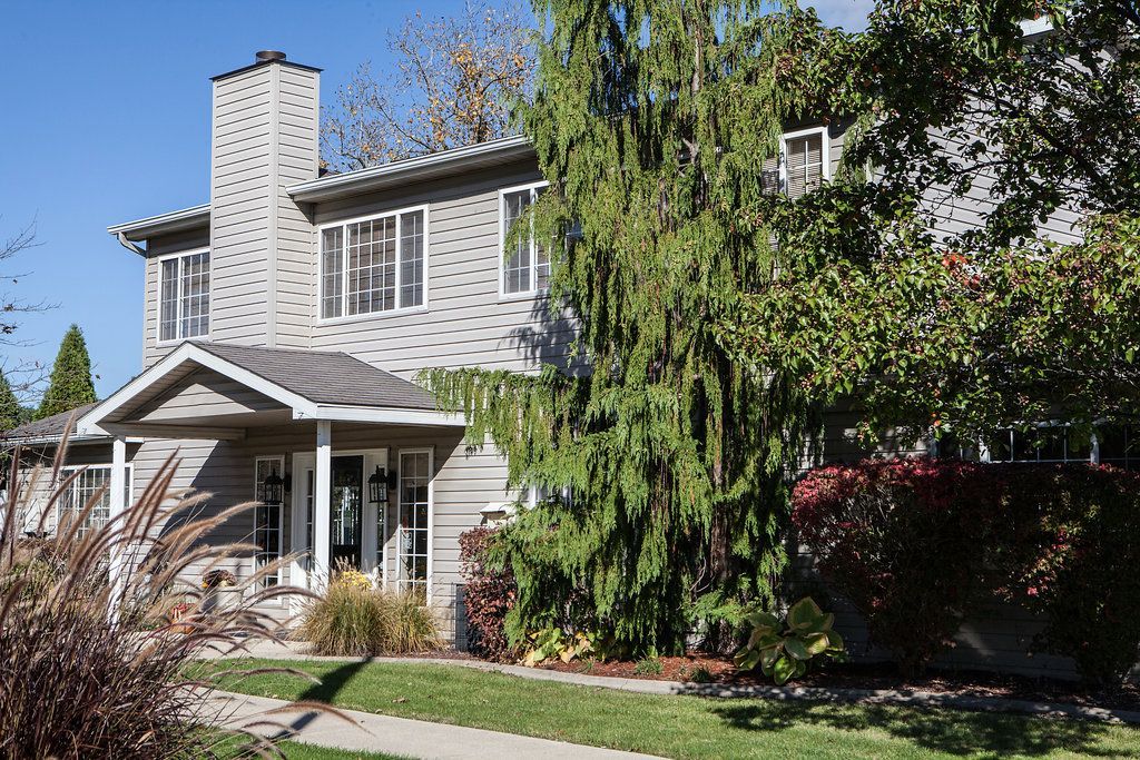 Two-story house with gray siding, a patterned chimney, and surrounding green trees under a blue sky.