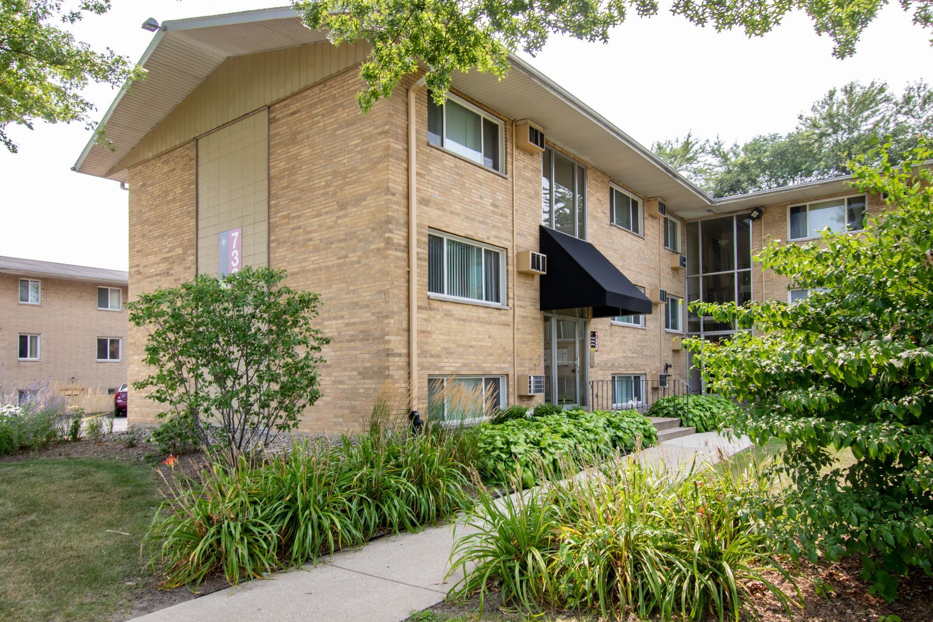 Apartment building with light brick facade, black awning, and landscaped front yard on a sunny day.