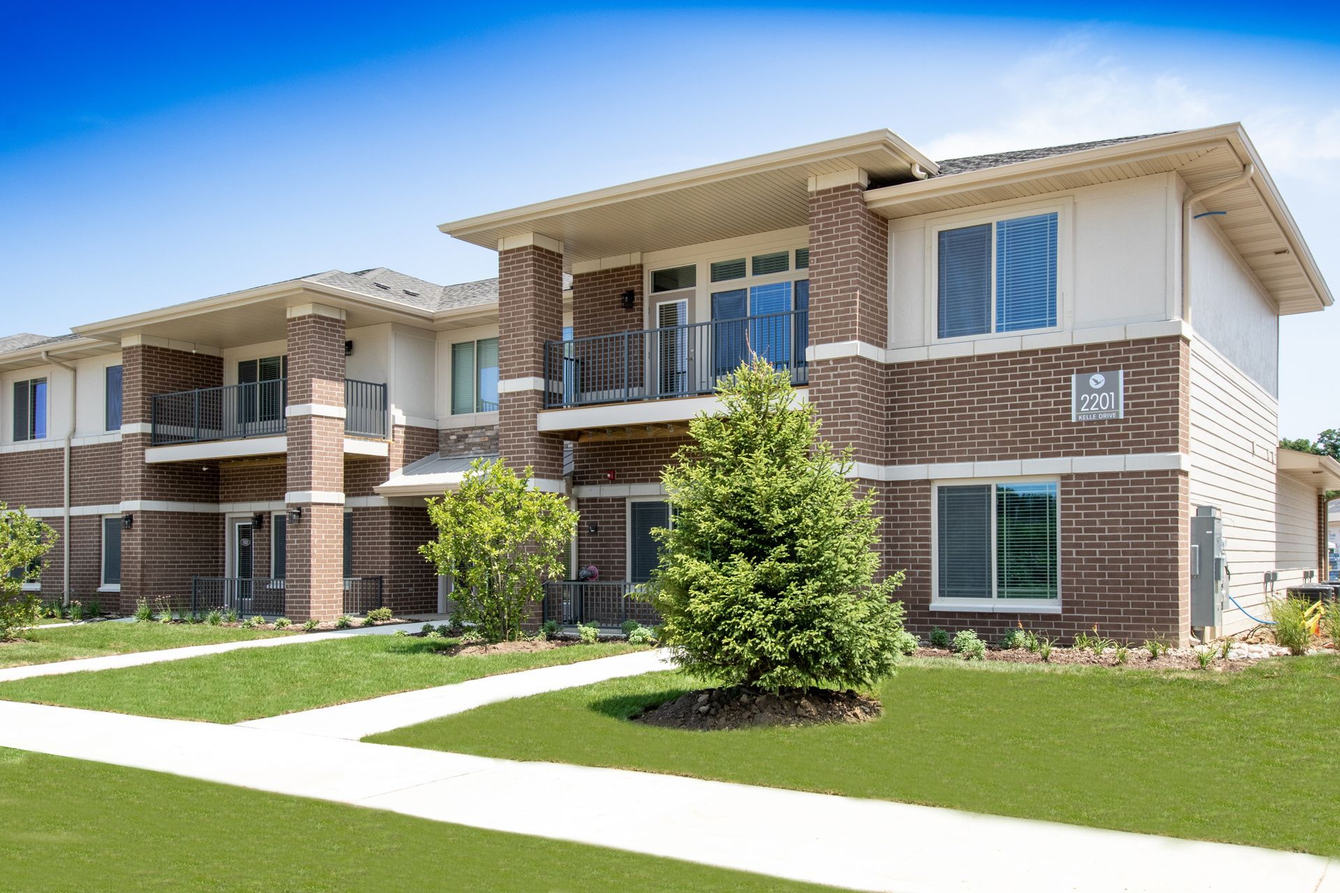 Two-story brick apartment building with balconies, windows, and green lawn under a blue sky.