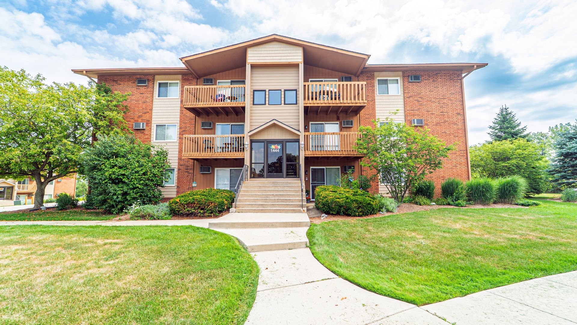 Three-story brick apartment building with brown trim and balconies; exterior stairs leading to central entrance.