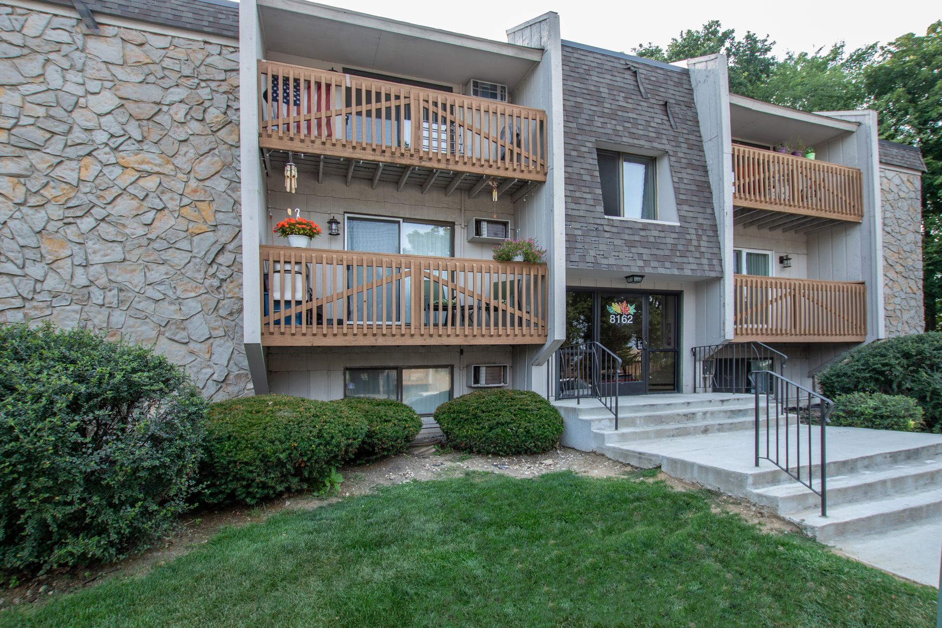 Three-story apartment building with stone facade, balconies, and entrance stairs. A car is parked on the left.