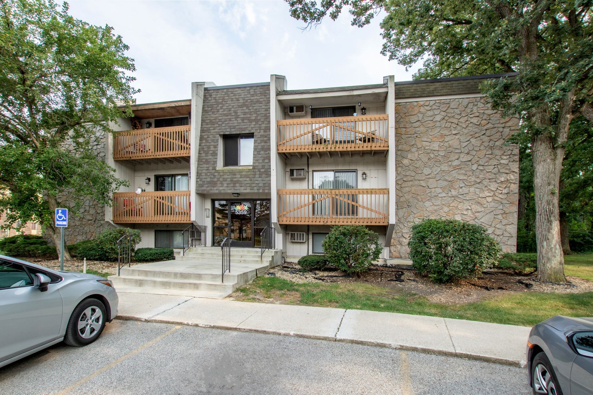 Apartment building with stone facade, balconies, and entrance stairs. Cars parked in front, trees on the side.