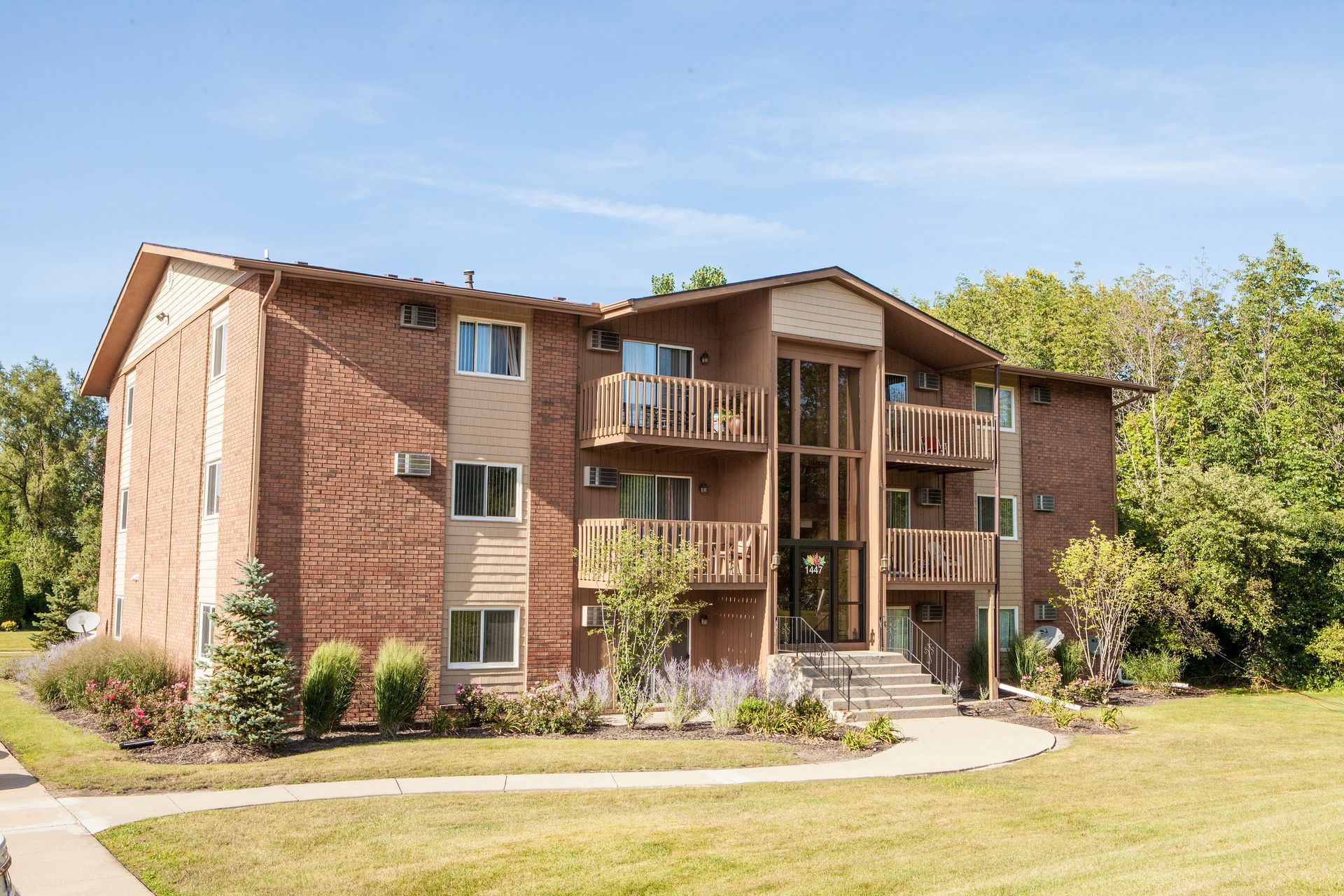 Three-story brick apartment building with balconies, steps, and green lawn under a blue sky.
