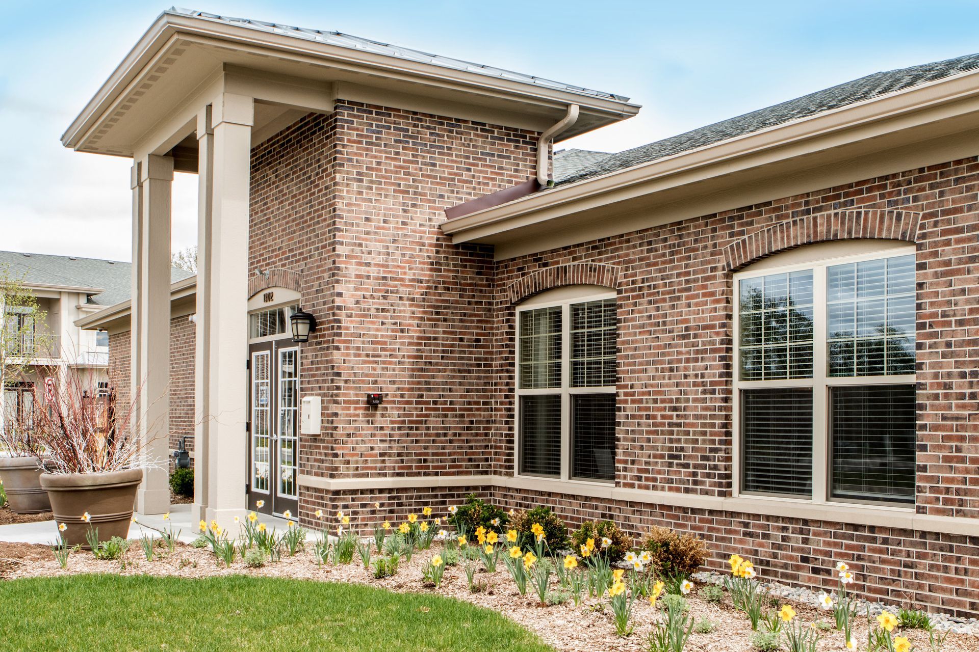 Brick building with arched windows and entryway under a portico, flower beds in front.