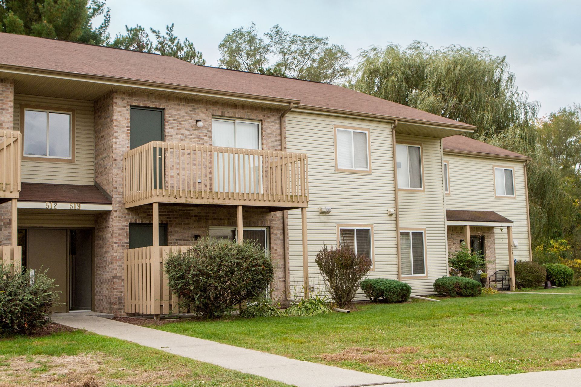Apartment building exterior, two-story with wooden decks, tan siding, and brick accents. Fall foliage in background.