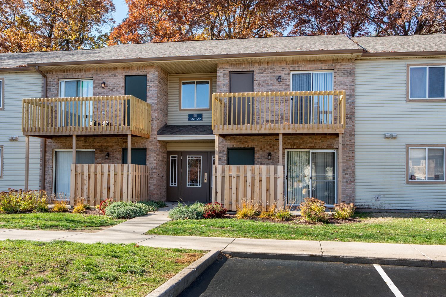 Two-story apartment building with balconies, wooden fences, and autumn trees in the background.