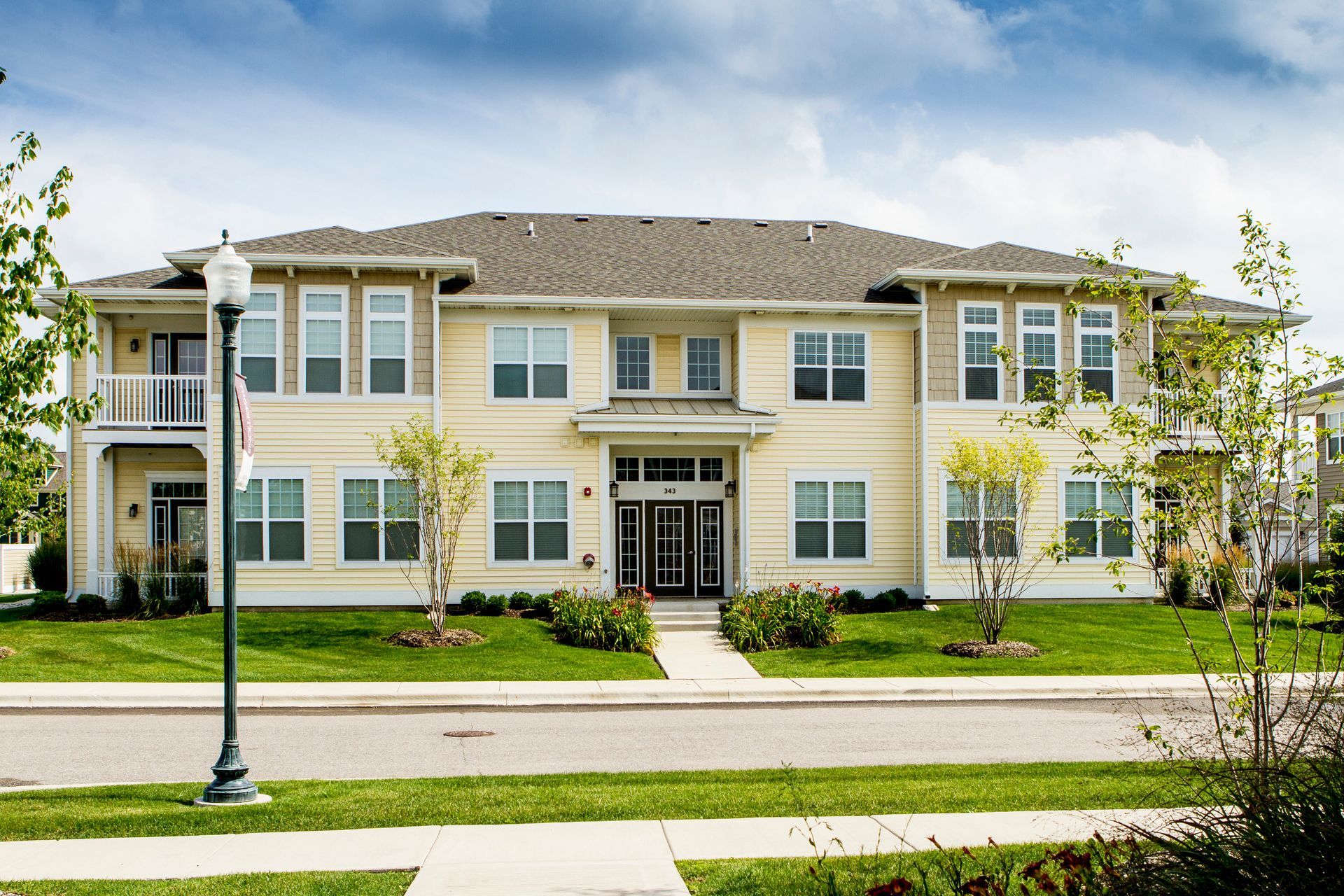Two-story apartment building with beige facade, green lawn, windows, and a dark shingled roof.