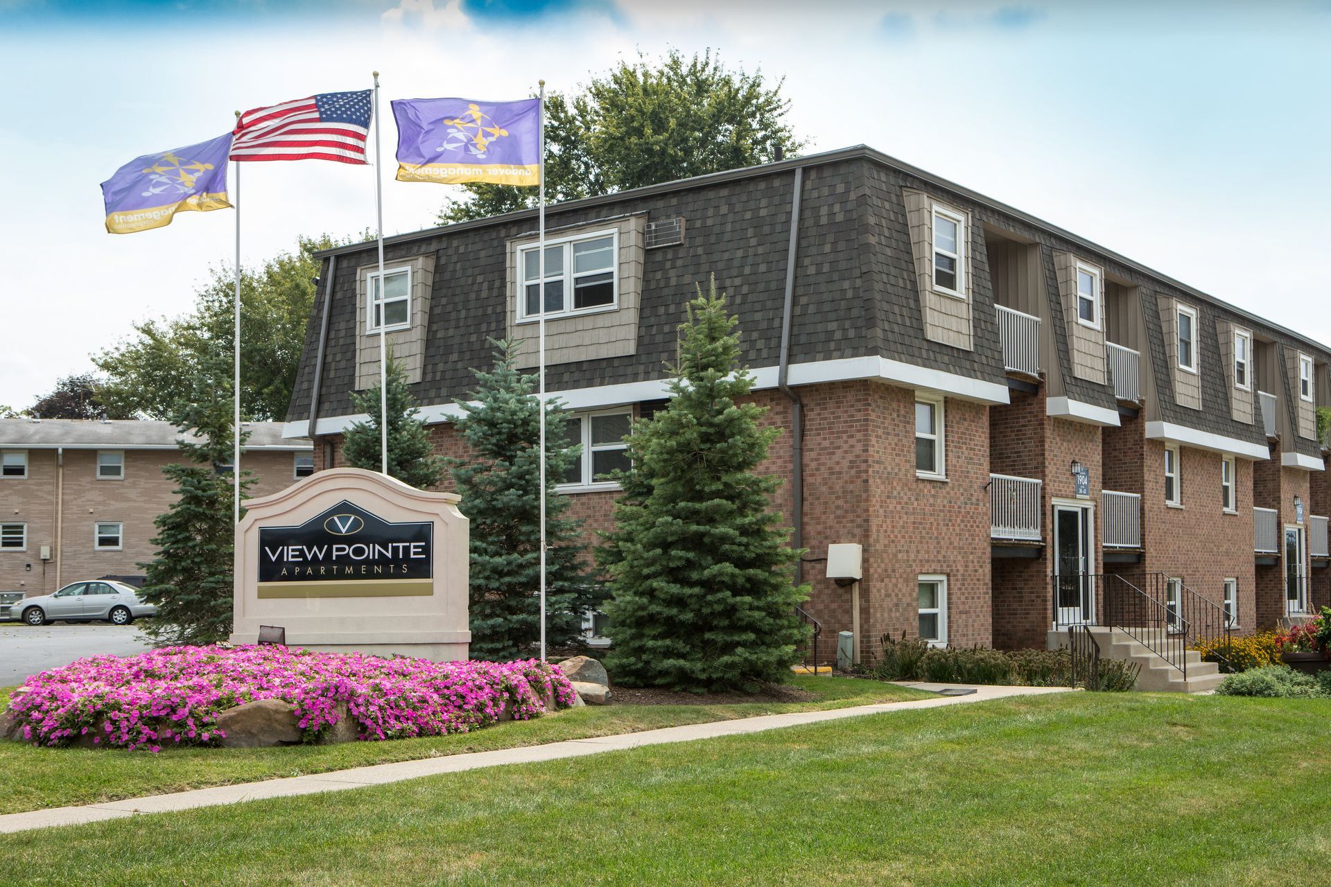 Apartment building with brick exterior, green lawn, flowerbed, and flags waving.