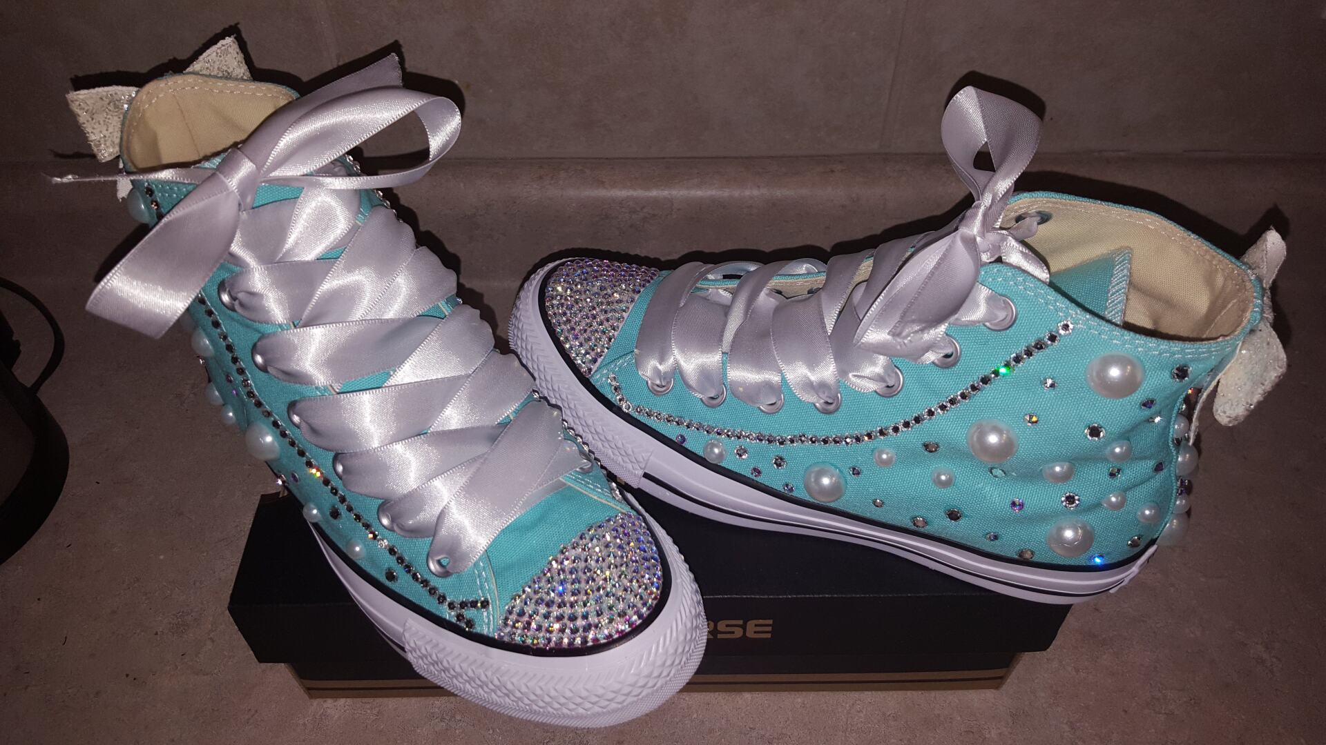 Turquoise high-top Converse sneakers embellished with rhinestones, pearls, and white ribbon laces, resting on a black box.