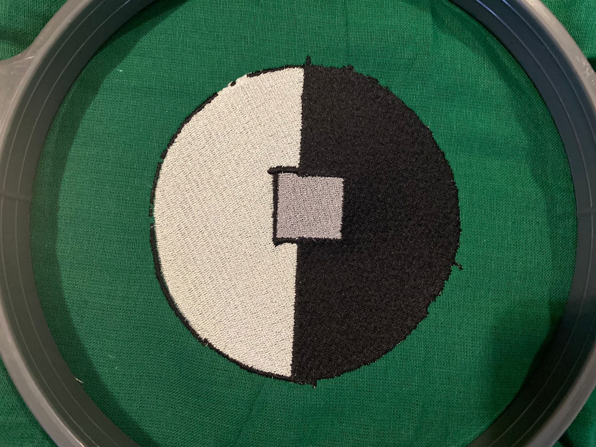 A black and white circle with a square in the middle is being embroidered on a green cloth.