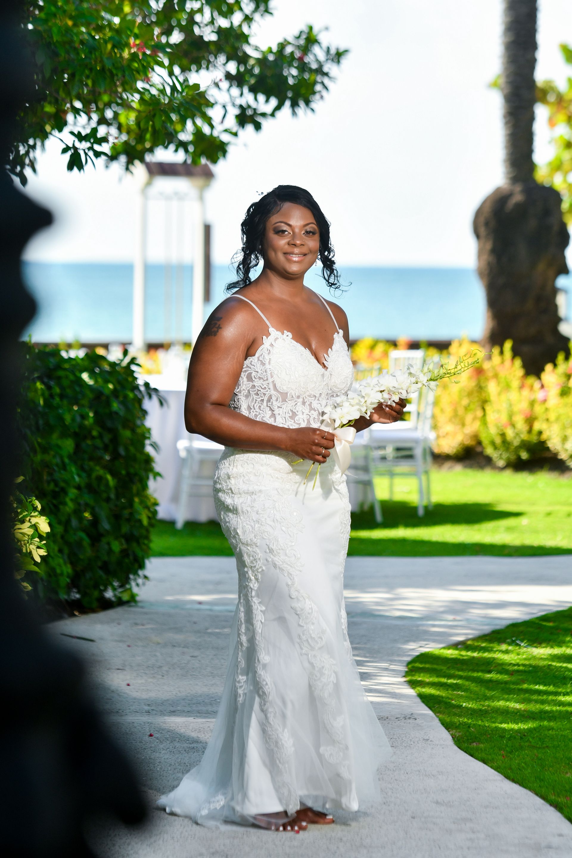 Bride in white lace gown holding bouquet on a garden path by the water