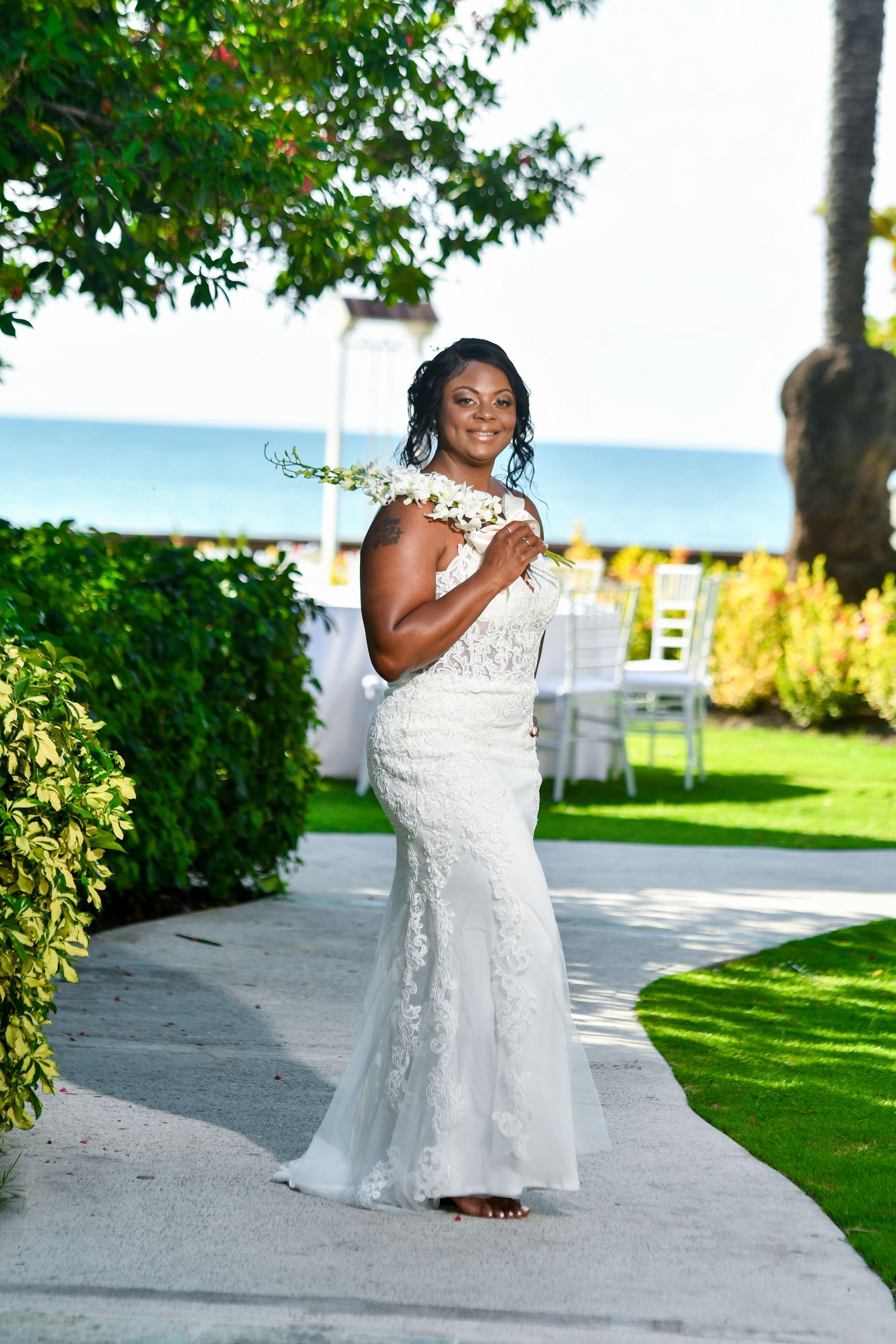 Woman in a white gown standing outdoors on a garden path by the water, smiling and holding a bouquet