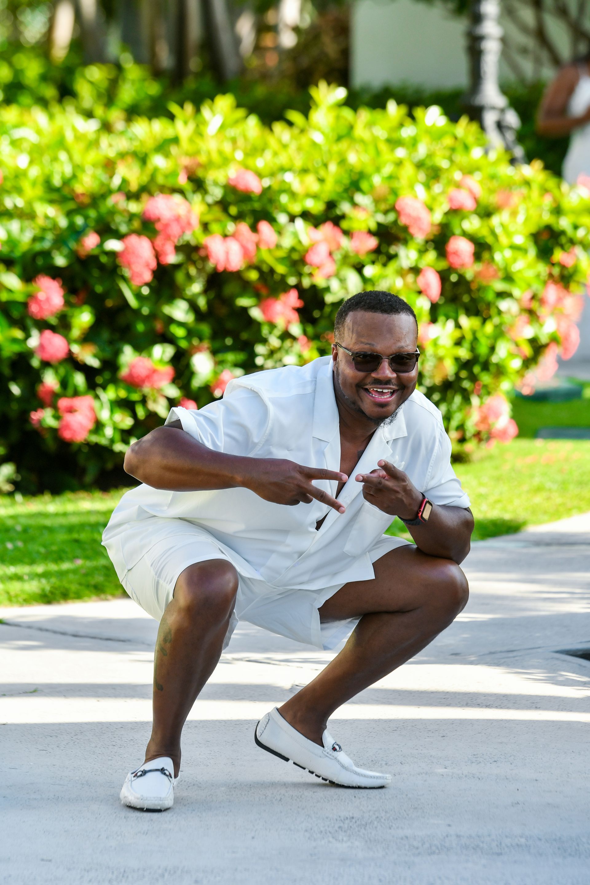 Man crouching on a sidewalk in a white outfit and sunglasses, smiling with lush green bushes behind him
