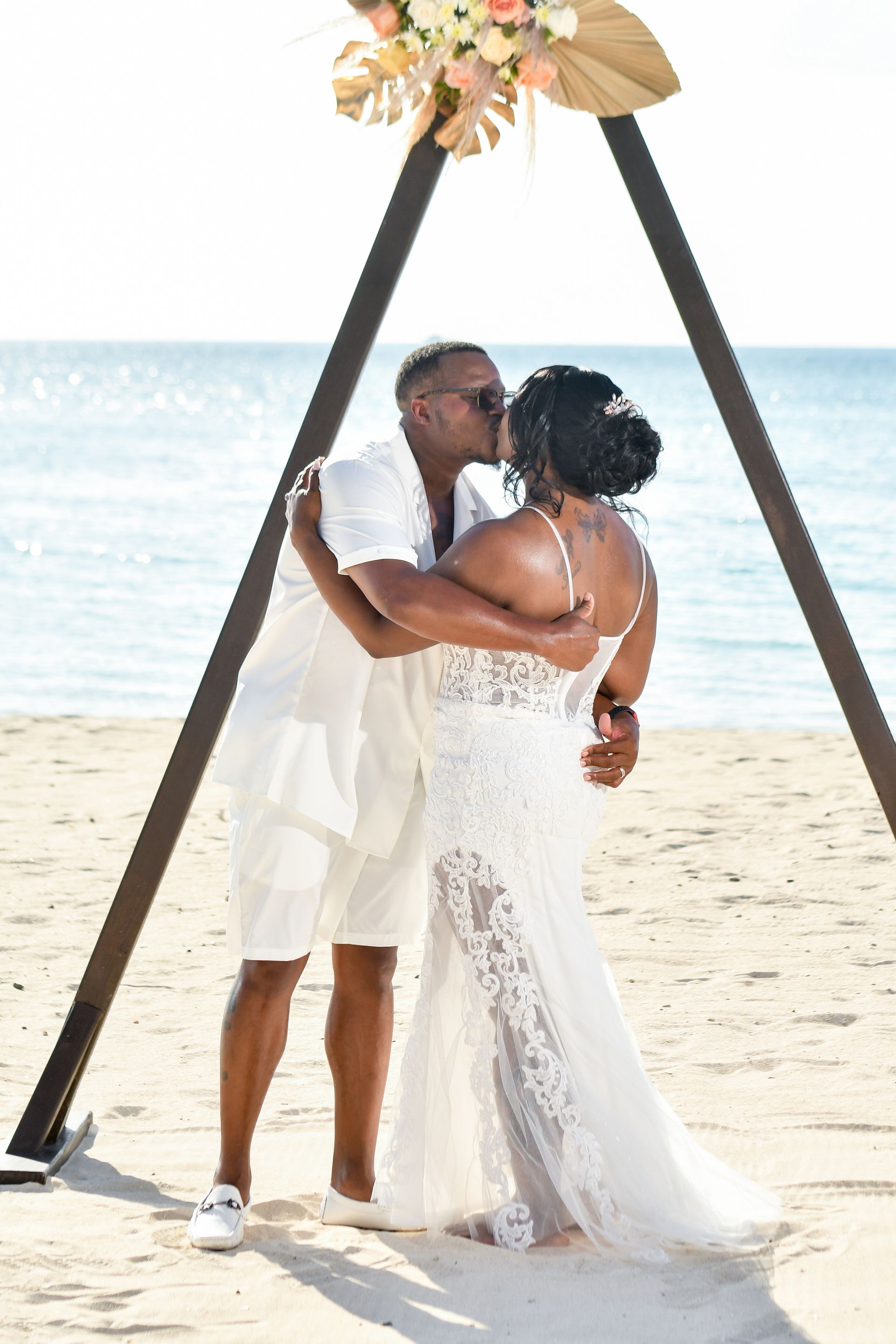 Couple in white attire kissing under a floral beach arch by the ocean.