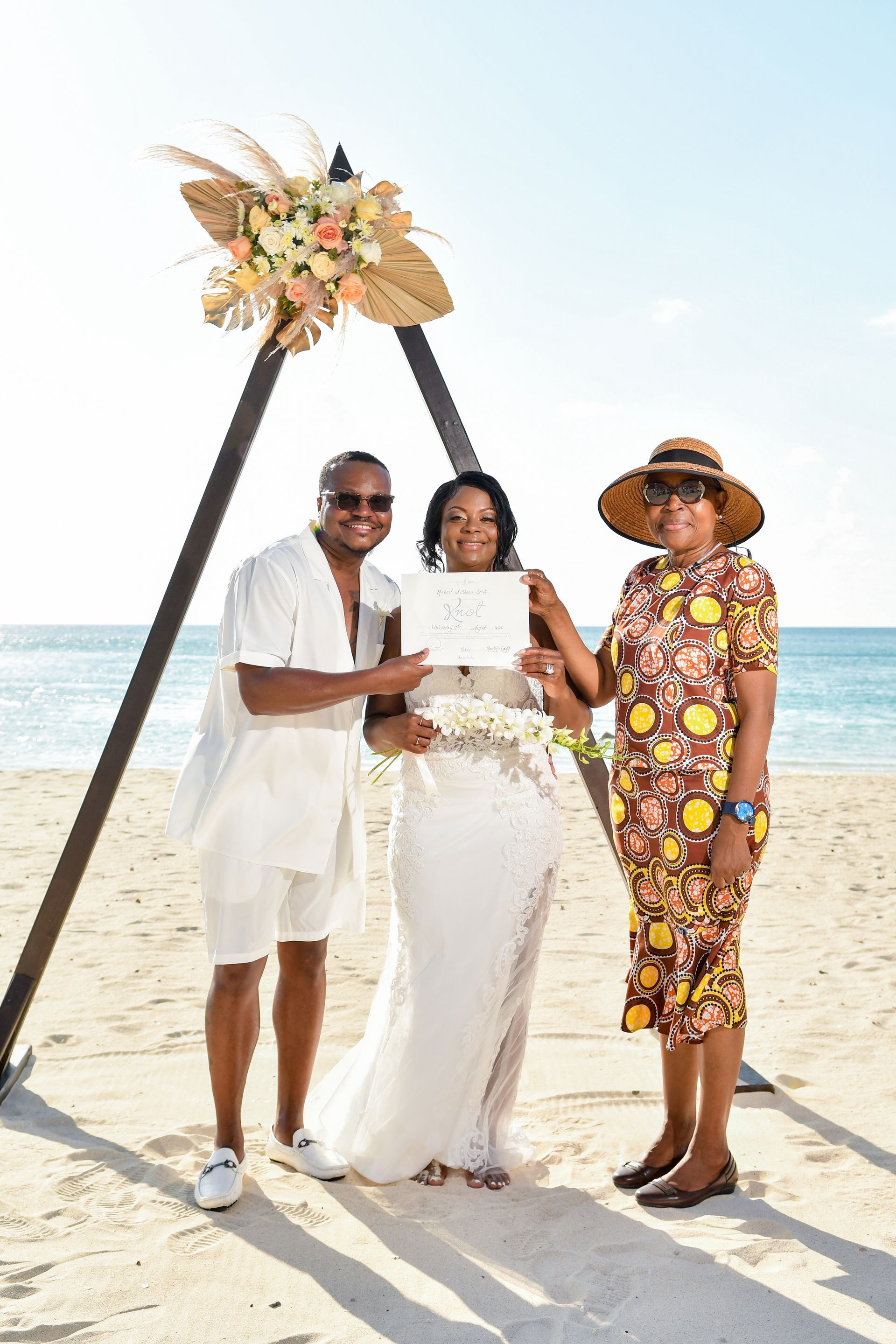 Three people in white and floral outfits pose on a beach beneath a floral arch.