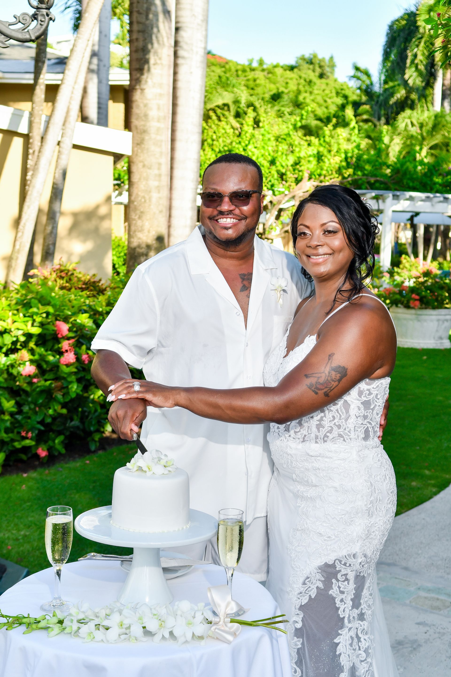 Couple in white wedding attire smiling beside a white cake outdoors on a garden path