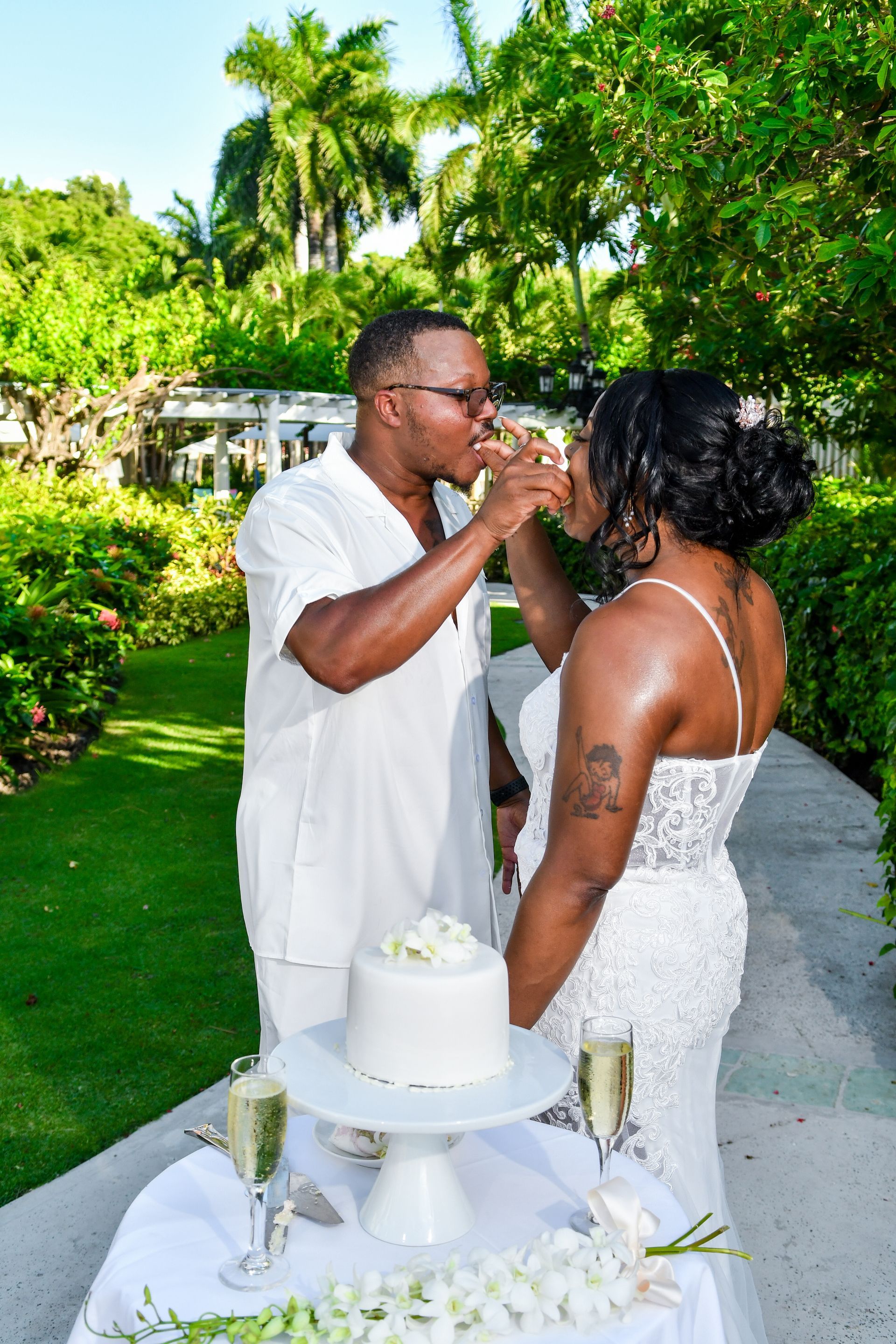 Couple feeding each other cake at an outdoor wedding table, with champagne and greenery around them