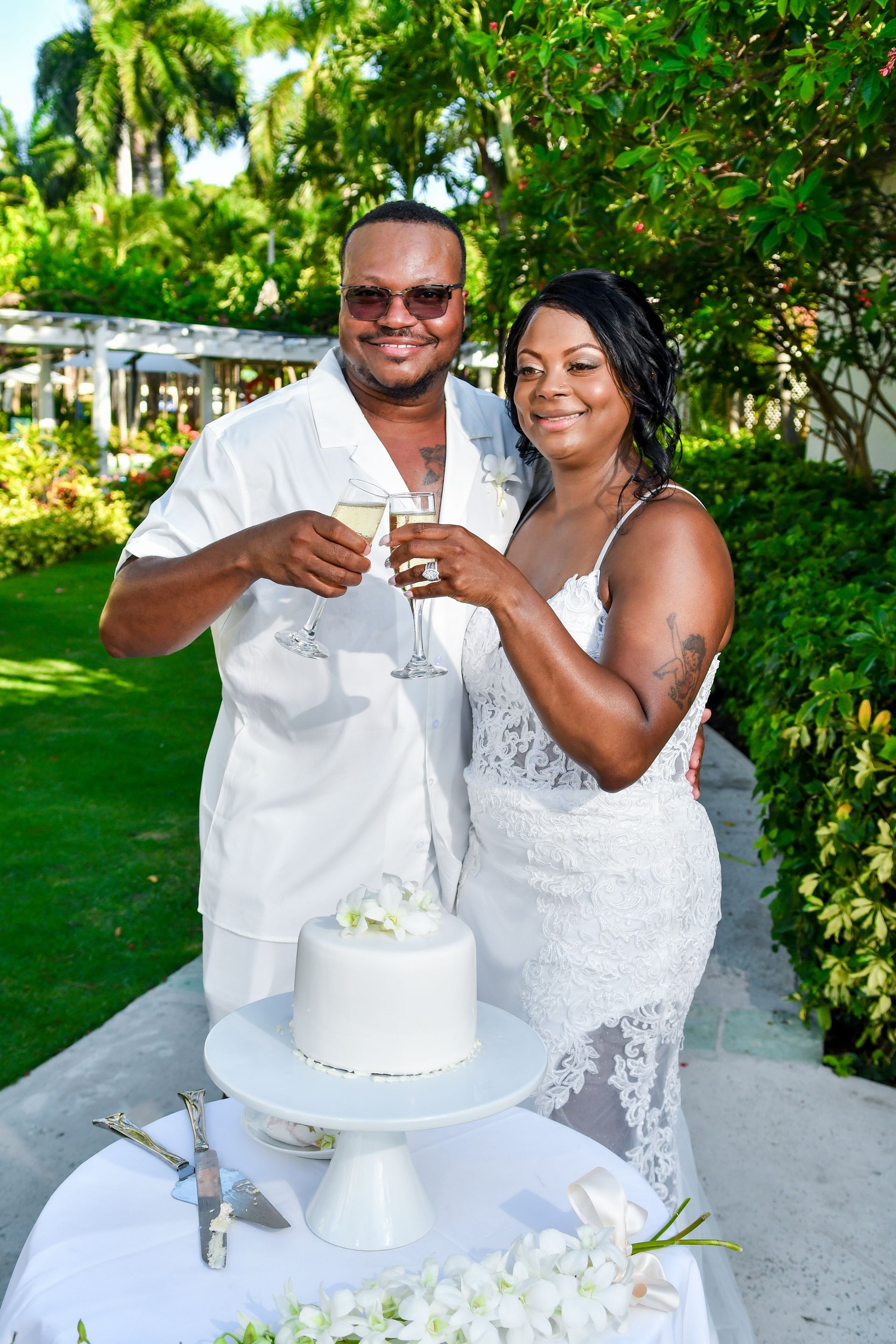 Couple toasting beside a white wedding cake outdoors in a lush garden