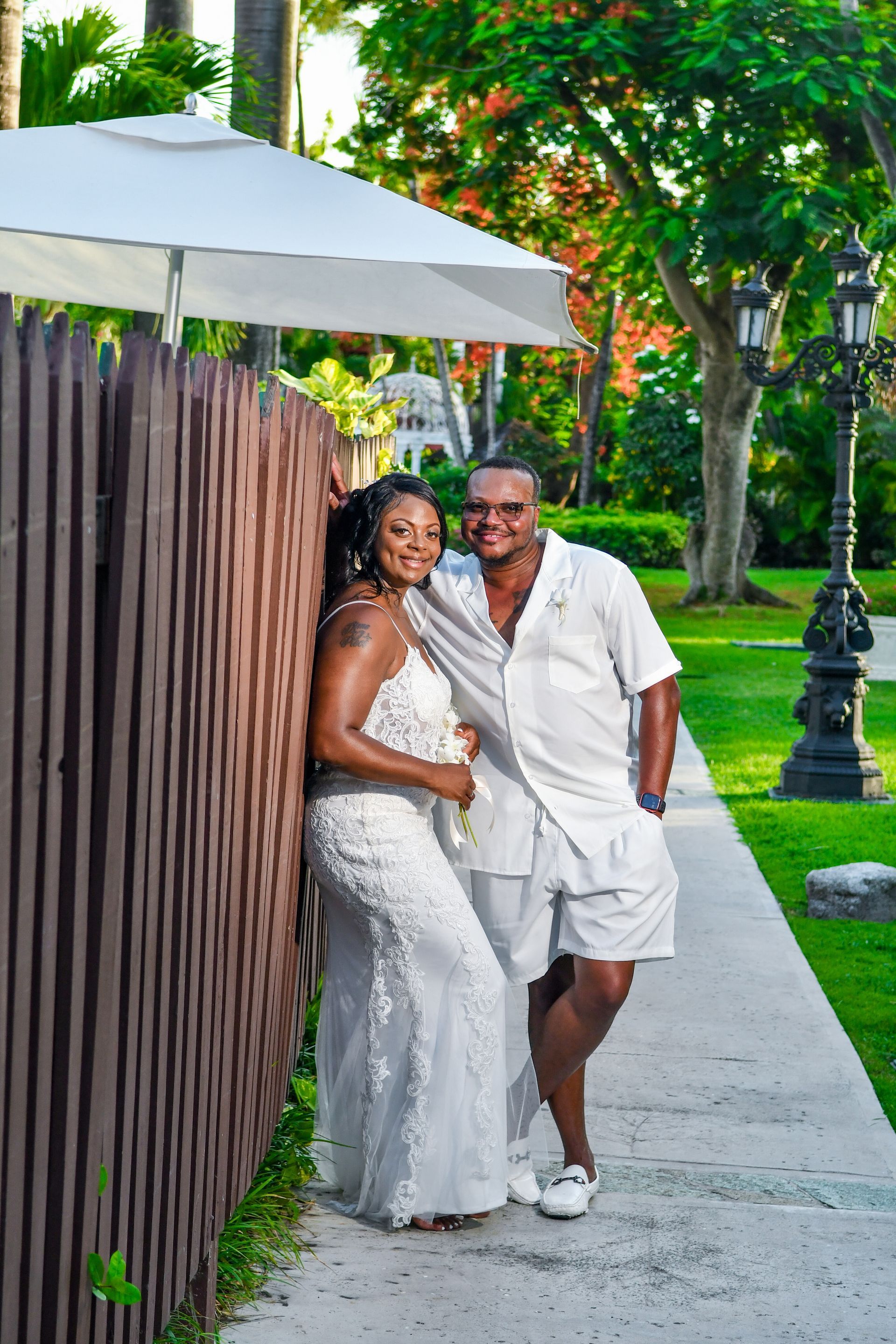 Couple in white outfits posing on a garden walkway beside a wooden fence and umbrella