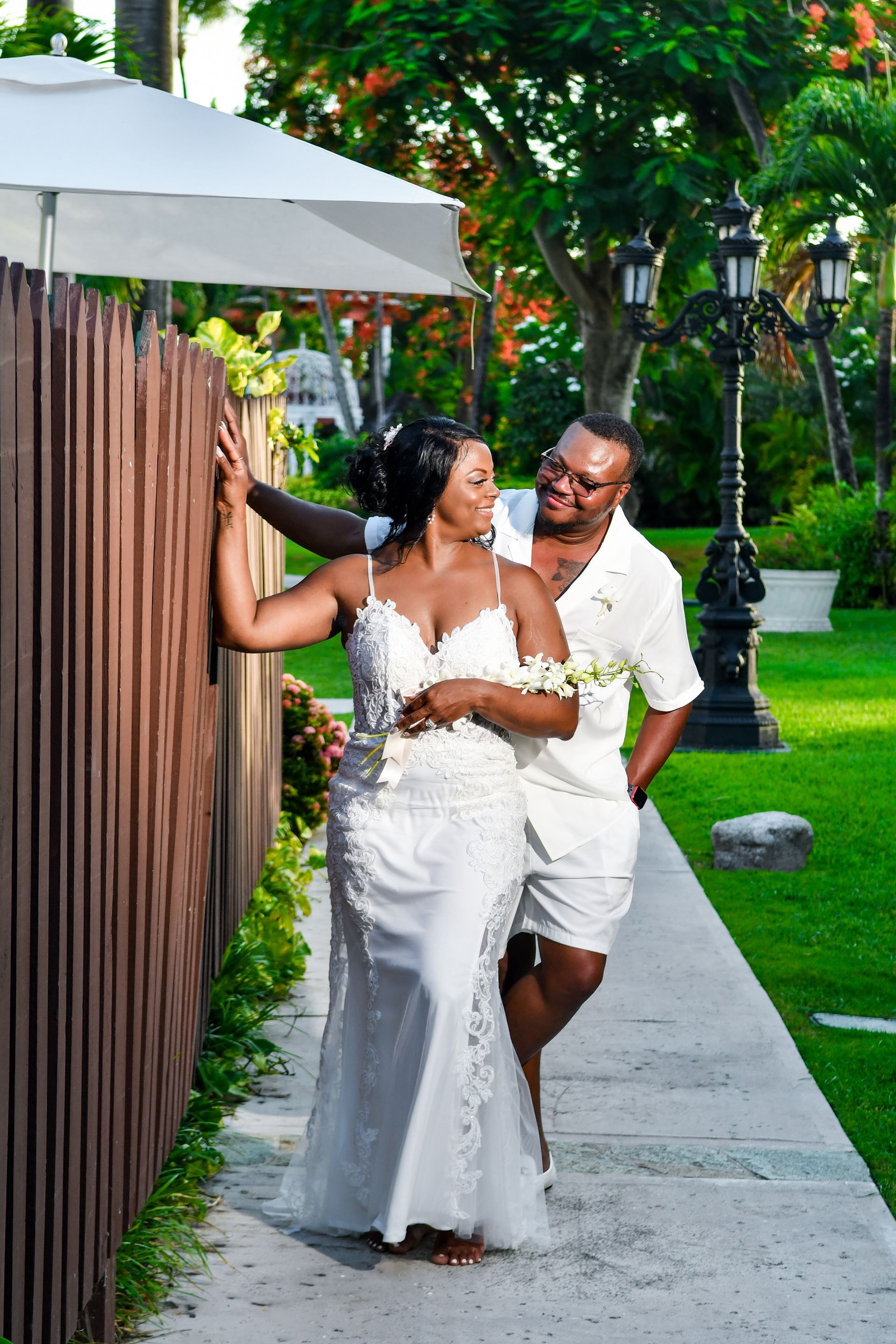 Couple in white wedding attire smiling on a garden path, with the bride holding a fence and a white umbrella overhead