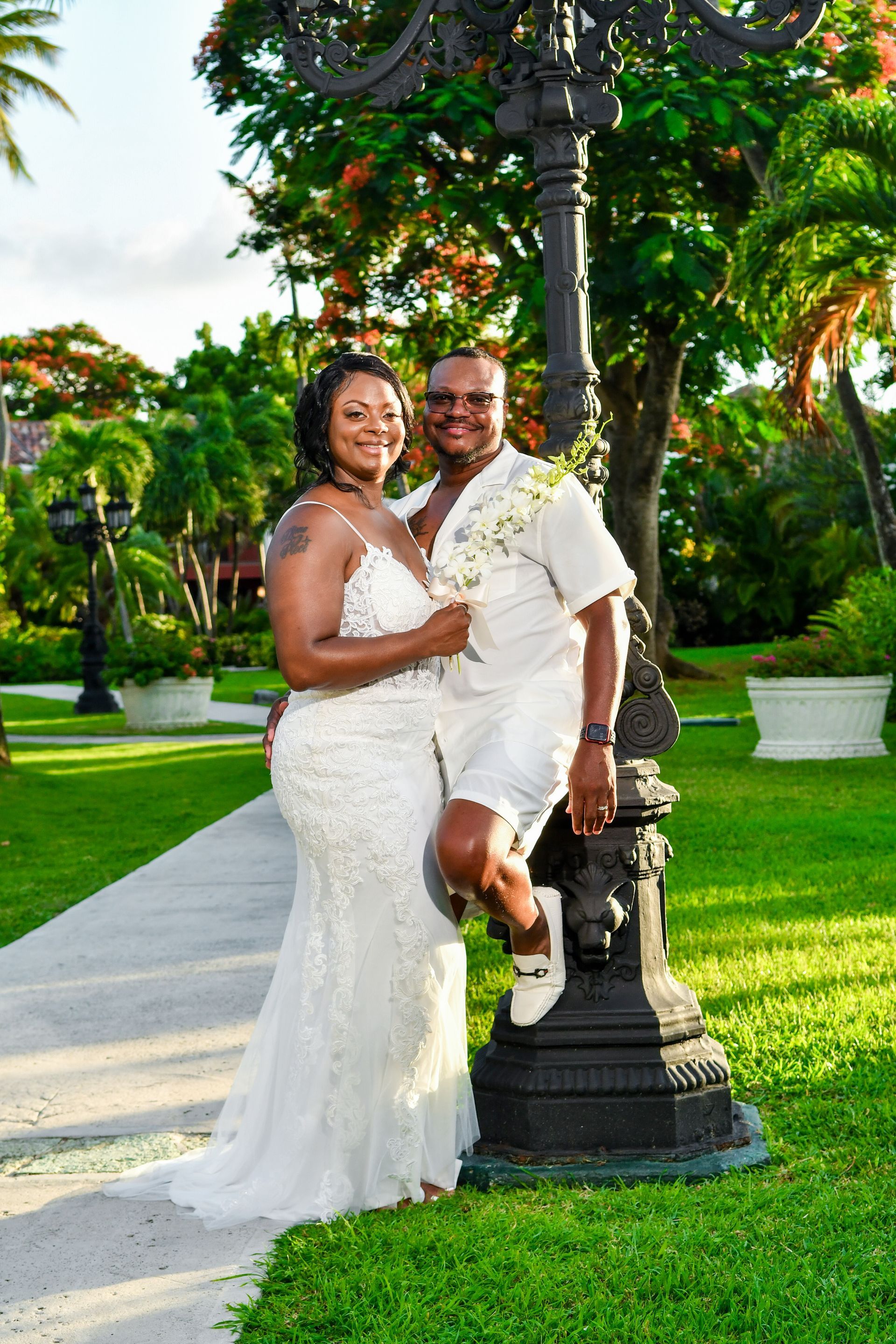 Couple in white wedding attire posing by a lamp post in a green garden walkway