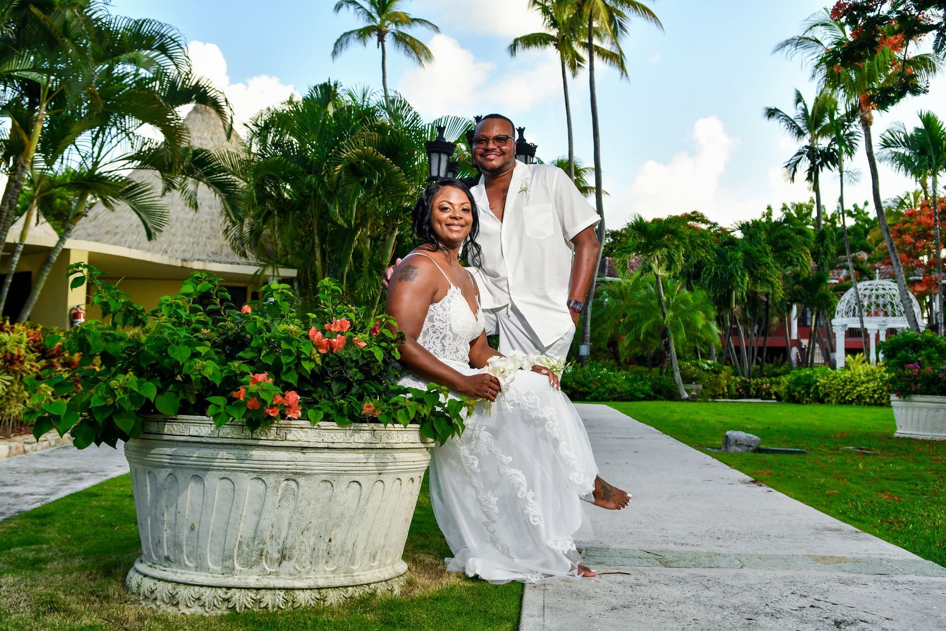 Couple in white standing on a garden path with palm trees and lush tropical landscaping.